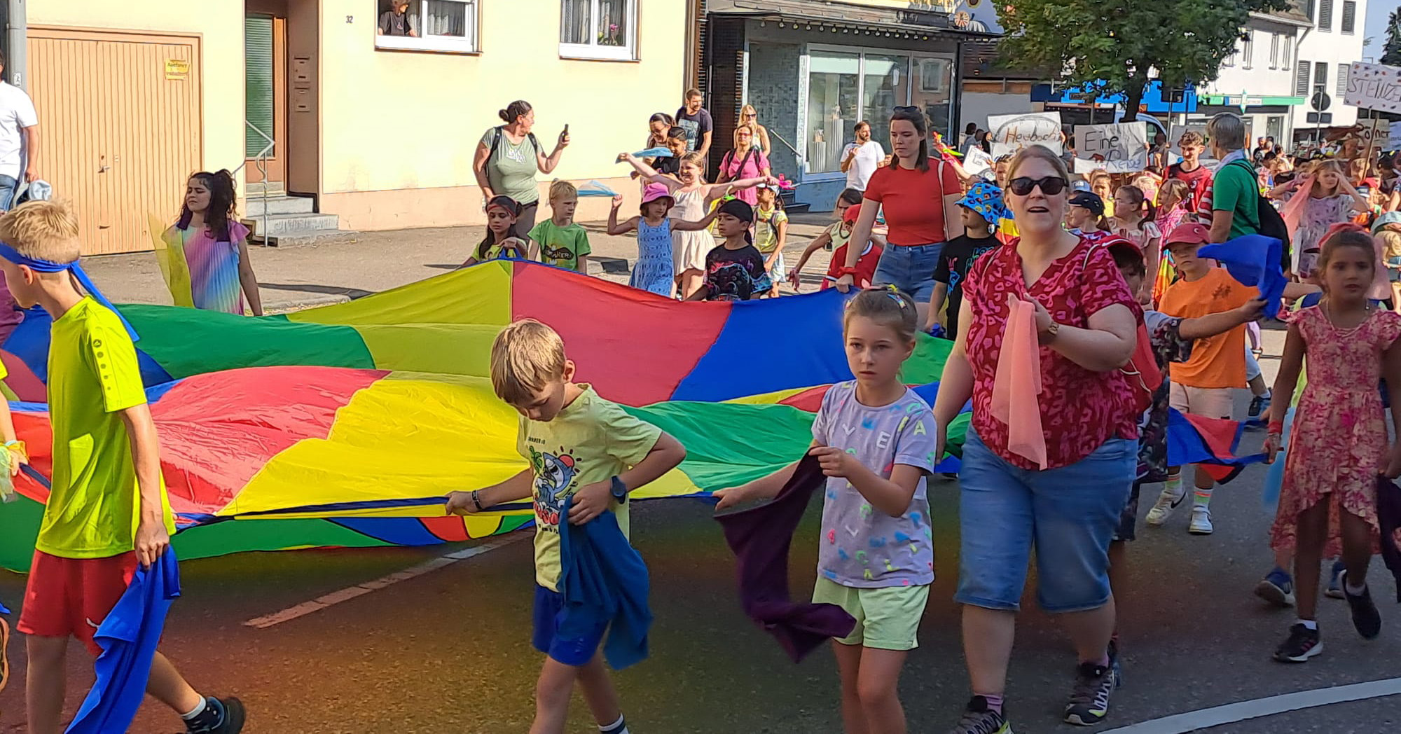 Eine Gruppe von Kindern und Erwachsenen läuft in einer farbenfrohen Parade, in der Hand einen großen Regenbogenfallschirm und Stoffteile. Sie sind draußen an einem sonnigen Tag, umgeben von Gebäuden und Zuschauern, und genießen die lebhafte Szene wie einen automatisch gespeicherten Entwurf.