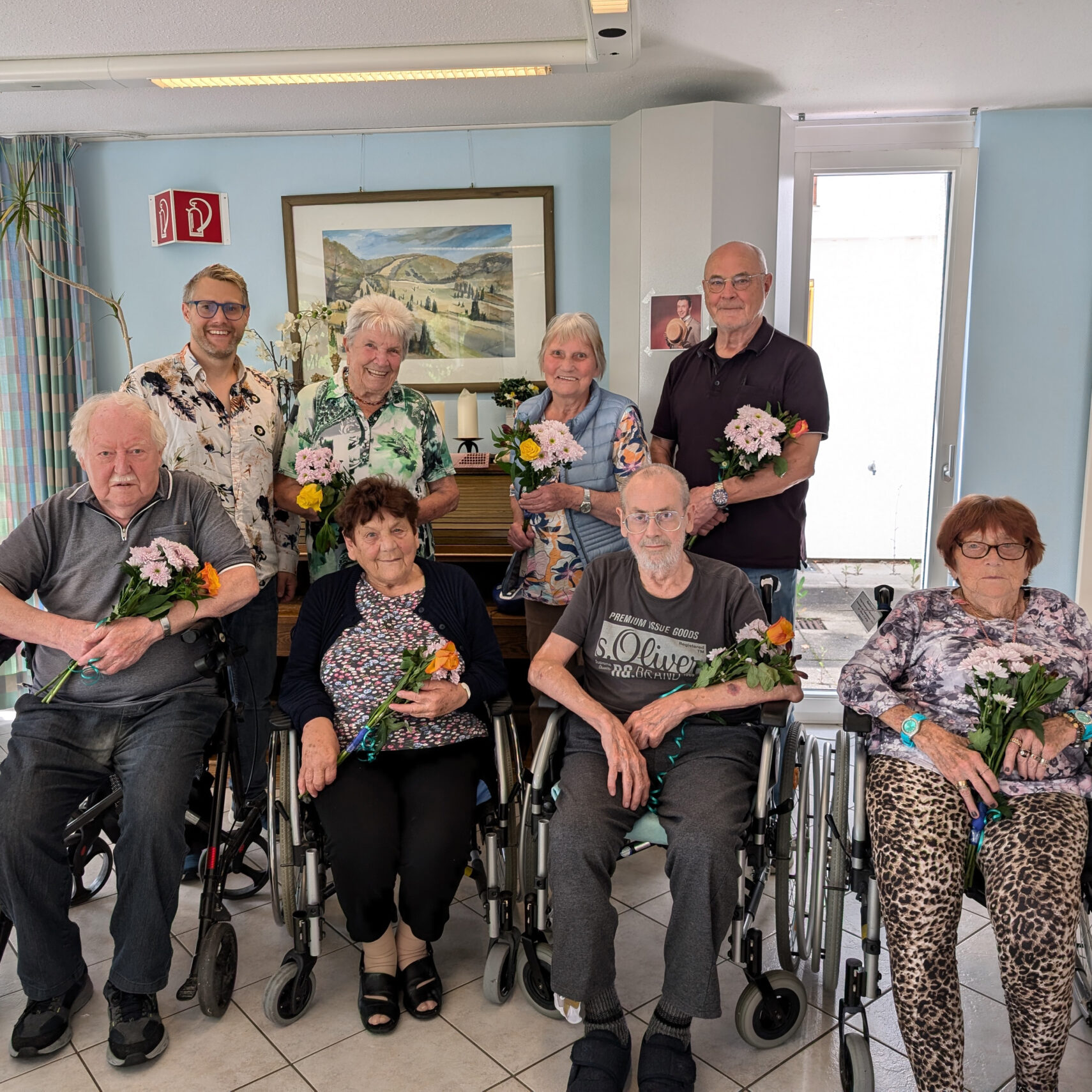 Eine Gruppe von acht lächelnden älteren Menschen, einige in Rollstühlen, hält Blumensträuße in einem Raum. Sie sind in einem hellen Raum mit Bildern und einem Klavier versammelt und feiern ihren neuen Heimbeirat im Sinne der Altenpflege.