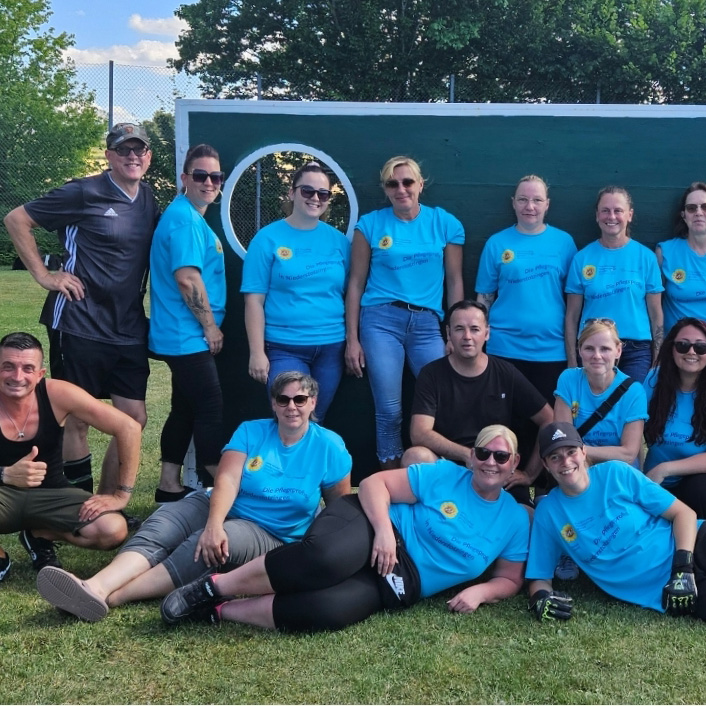 Eine Gruppe von Menschen, meist in passenden blauen T-Shirts, posiert gemeinsam im Gras vor einer grünen Tafel mit weißen Kreisen - wie ein automatisch gespeicherter Entwurf - im Sonnenschein. Im Hintergrund sind Bäume und ein Zaun zu sehen.