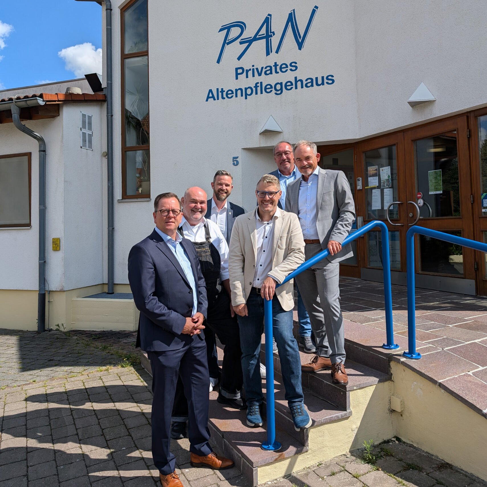 Sechs Männer in Geschäftskleidung stehen an einem sonnigen Tag lächelnd auf den Stufen vor einem Gebäude mit der Aufschrift PAN Privates Altenpflegehaus, einem Partner der Stiftung Haus Lindenhof.