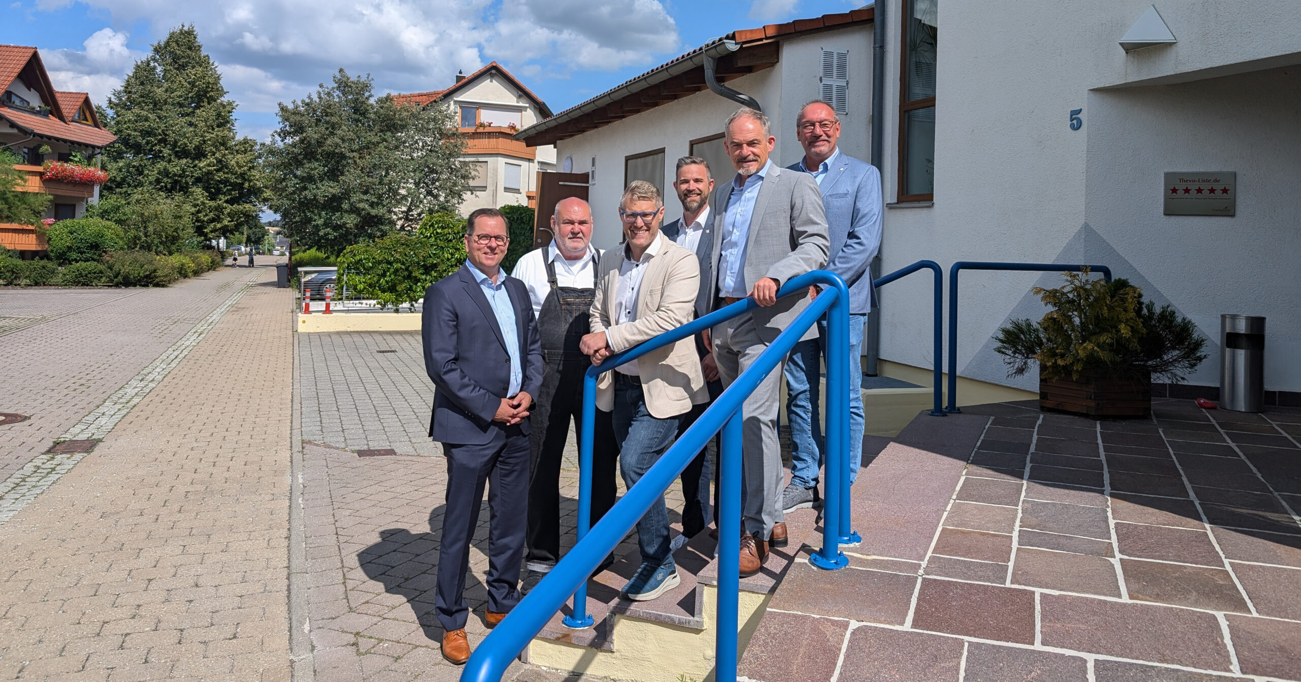 Sechs Männer in Geschäftskleidung stehen gemeinsam auf einer Treppe mit blauem Geländer vor der Stiftung Haus Lindenhof und posieren an einem sonnigen Tag für ein Gruppenfoto. Im Hintergrund sind Wohnhäuser und Grünanlagen in St. Helena zu sehen.