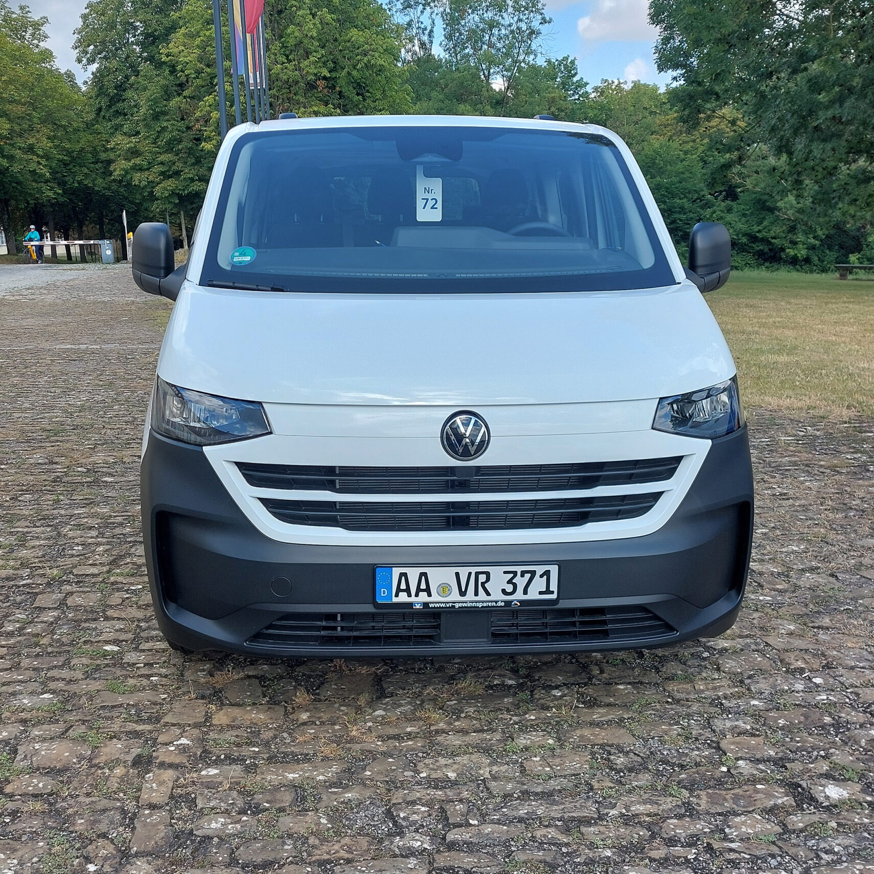 Ein weißer Volkswagen VR-Bus ist auf einem Kopfsteinpflaster im Freien in Strümpfelbach geparkt, umgeben von grünen Bäumen. Der Bus hat ein deutsches Kennzeichen AA VR 371 und an der Windschutzscheibe ist ein Zettel mit der Nummer 72 zu sehen.