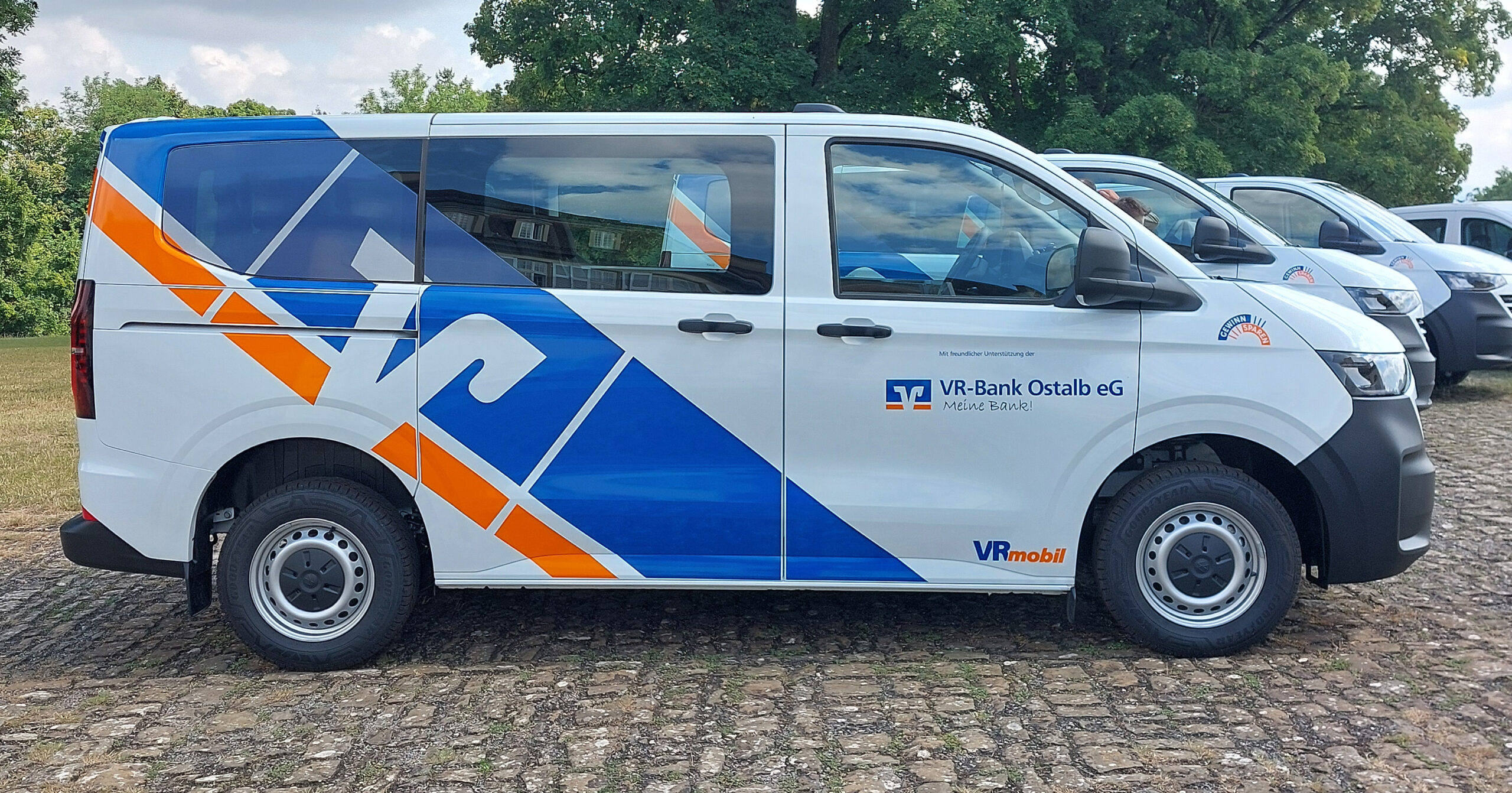 Ein weißer VR-Bus mit blauen und orangefarbenen geometrischen Motiven und dem Logo der VR-Bank Ostalb eG parkt auf einer gepflasterten Fläche mit Gras und Bäumen im Hintergrund. Weitere Fahrzeuge von Neuer Bus der WG Strümpfelbach sind in der Nähe geparkt.
