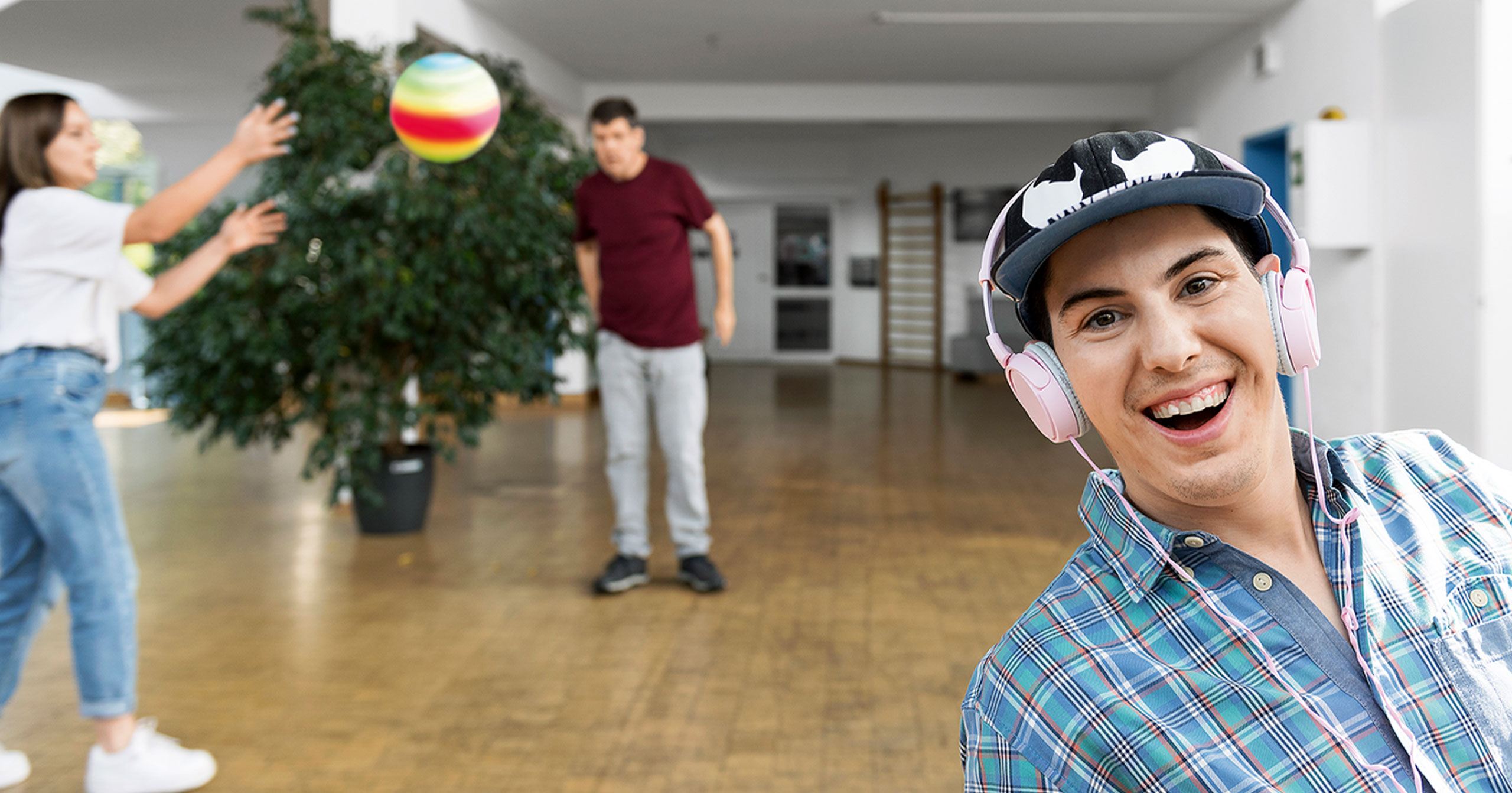 Im Vordergrund ist eine lächelnde Person mit Mütze und rosa Kopfhörern zu sehen, während sich zwei Personen im Hintergrund in einem geräumigen Innenraum mit Holzboden und einer großen Pflanze einen bunten Ball zuwerfen.