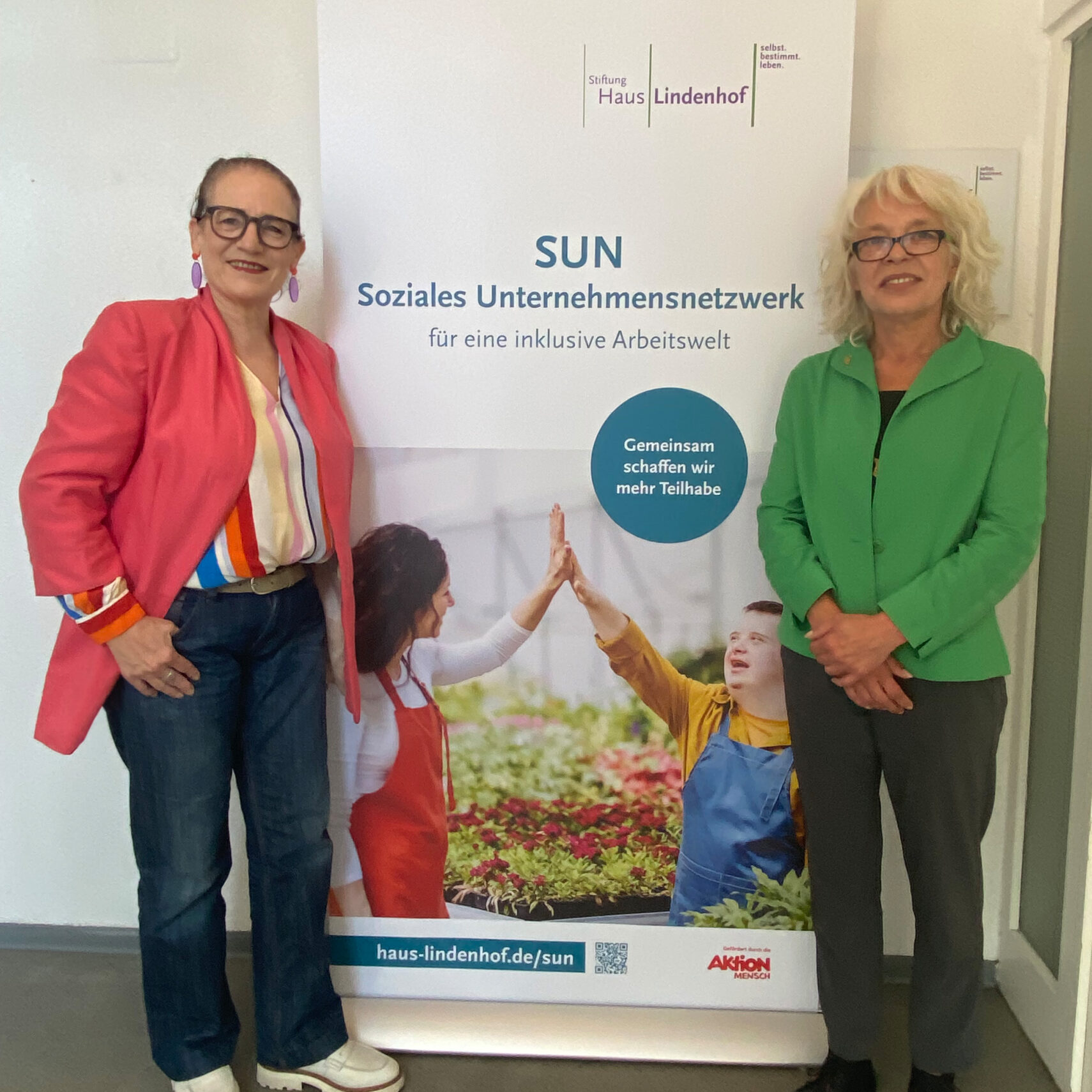 Zwei Frauen stehen lächelnd vor einem Banner des SUN Soziales Unternehmensnetzwerks, das mit dem Kompetenzzentrum Arbeit für inklusive Arbeitsplätze wirbt. Das Banner zeigt zwei Menschen in einem Gewächshaus und Informationen über das Programm von Martina Häusler.