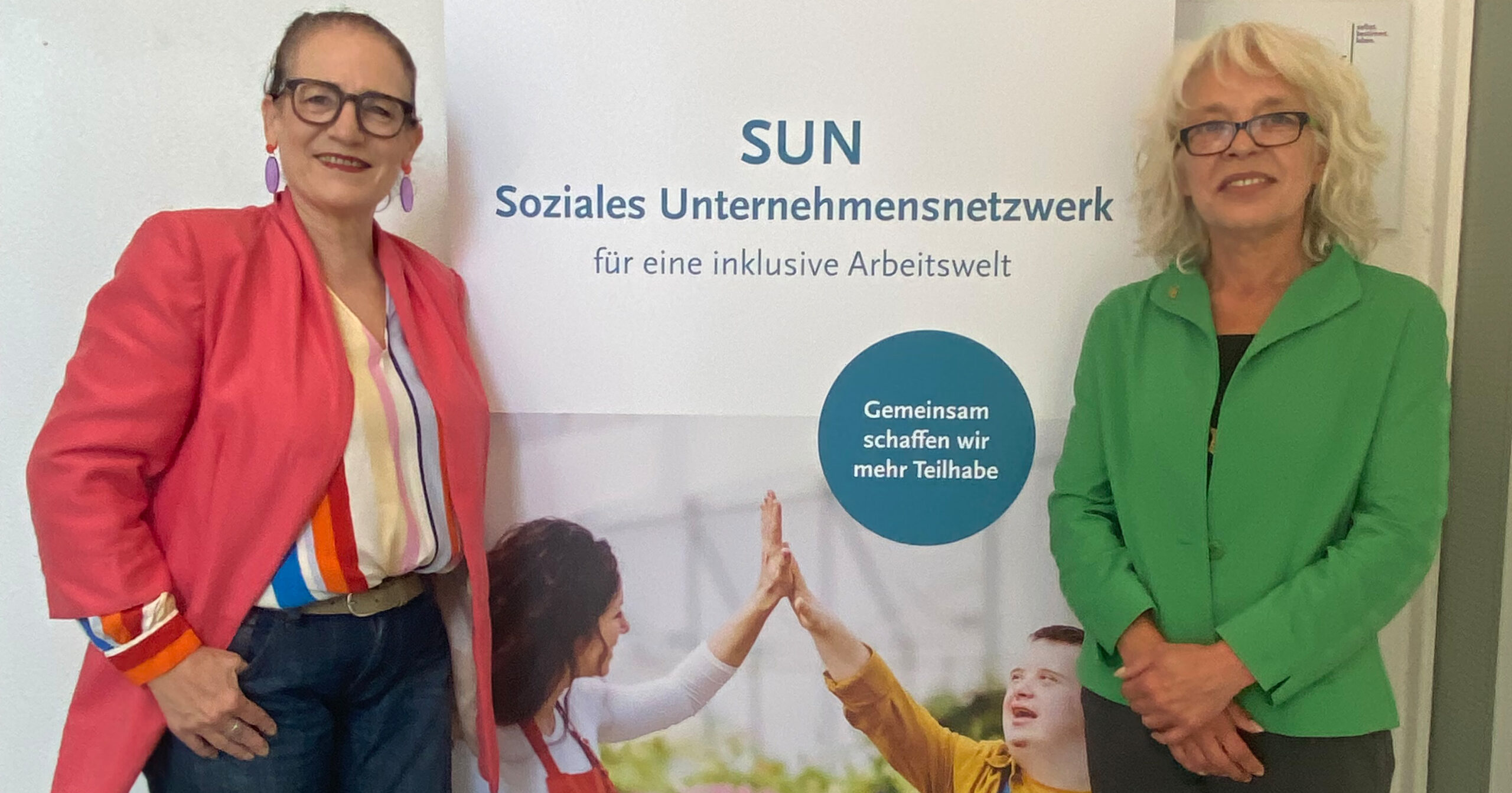 Zwei Frauen, darunter Martina Häusler, stehen lächelnd neben einem Schild für das SUN Soziales Unternehmensnetzwerk, auf dem Menschen sich gegenseitig zujubeln und die Botschaft Gemeinsam schaffen wir mehr Teilhabe zu lesen ist - eine Mission, die auch vom Kompetenzzentrum Arbeit unterstützt wird.