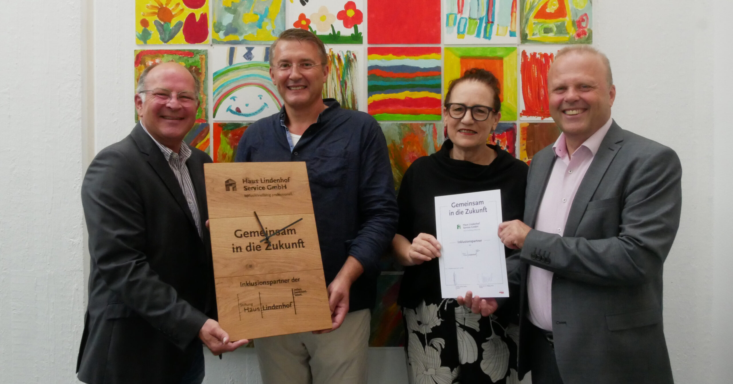 Vier Personen stehen lächelnd vor einem bunten Kunstwerk. Zwei von ihnen halten eine große Holztafel mit der Aufschrift Gemeinsam in die Zukunft für das Projekt SUN, während die andere Person, die HLS repräsentiert, eine Urkunde mit dem gleichen Titel in der Hand hält.