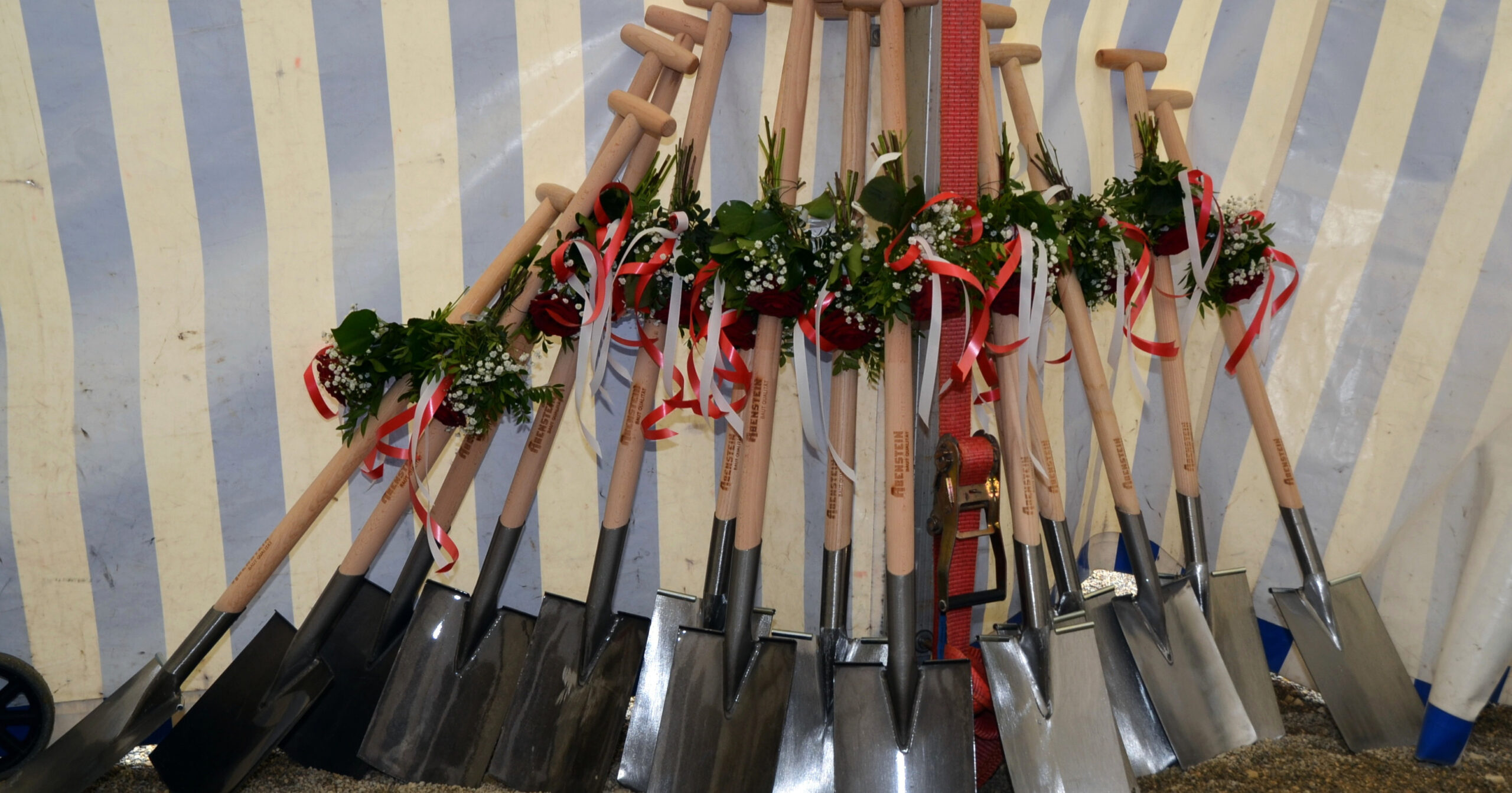 Eine Gruppe glänzender Schaufeln mit Holzgriffen, die mit roten und weißen Bändern und kleinen Blumensträußen geschmückt sind, lehnt an einer weiß-blau gestreiften Wand, bereit für die St. Helena Spatenstich-Zeremonie.