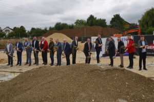 Eine Gruppe von fünfzehn Personen in förmlicher Kleidung steht in einer Reihe auf einer Baustelle, jeder hält eine Schaufel mit Erde in der Hand und posiert für einen automatisch gespeicherten Entwurf einer Grundsteinlegung. Im Hintergrund sind Schotterhaufen und Baumaschinen zu sehen.