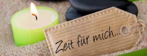 Eine grüne Kerze brennt im Sand neben einem Pappschild mit der Aufschrift "Zeit für mich..." - eine friedliche Erinnerung an Entspannung und sanftes Stressmanagement.