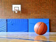 Ein Basketball liegt auf dem Turnhallenboden vor blau gepolsterten Wänden, bereit für das nächste Training der Basketballgruppe unter einem an einer Ziegelwand befestigten Korb.