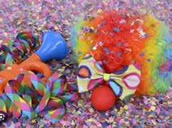 Auf einer mit Konfetti bedeckten Fläche liegen Clown-Accessoires, die sich perfekt für Karneval oder Fasching eignen, darunter eine lockige Regenbogenperücke, eine rote Nase, eine bunte Fliege und ein Geräuschmacher.