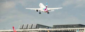 Ein Verkehrsflugzeug hebt über dem Flughafenterminal ab, während andere Flugzeuge und Flughafengebäude am Boden unter dem bewölkten Himmel zu sehen sind.