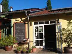Ein sonniges Hofcafé im Freien mit gelben Wänden, weiß umrahmten Türen, einer offenen Tür, Topfpflanzen und einer großen Speisekarte auf einer Kreidetafel am Eingang - der perfekte Ort für einen entspannten Ausflug.