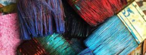Eine Nahaufnahme von mehreren gebrauchten Pinseln mit Borsten, die in verschiedenen Farben gefärbt sind, darunter blau, rot, schwarz und hellblau, die auf einer strukturierten Oberfläche liegen.