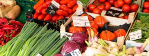 An einem farbenfrohen Marktstand wird frisches Gemüse, darunter Tomaten, Frühlingszwiebeln, Rotkohl, Knoblauch, Chilischoten und kleine orangefarbene Kürbisse, mit handgeschriebenen Preisschildern angeboten.
