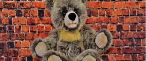 Ein grauer Teddybär sitzt vor einem roten Backsteinhintergrund und blickt mit ausgestreckten Armen und freundlichem Gesichtsausdruck nach vorne - genau wie die Klassiker, die Sie im Steiff Museum, der Heimat der legendären Steiff-Teddybären, finden.