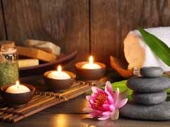 Drei brennende Kerzen, gestapelte Massagesteine, eine rosafarbene Lotusblume, ein aufgerolltes weißes Handtuch und Spa-Accessoires sind auf einer hölzernen Fläche neben einem Wasserklangbett angeordnet und vermitteln eine beruhigende und entspannende Spa-Atmosphäre.