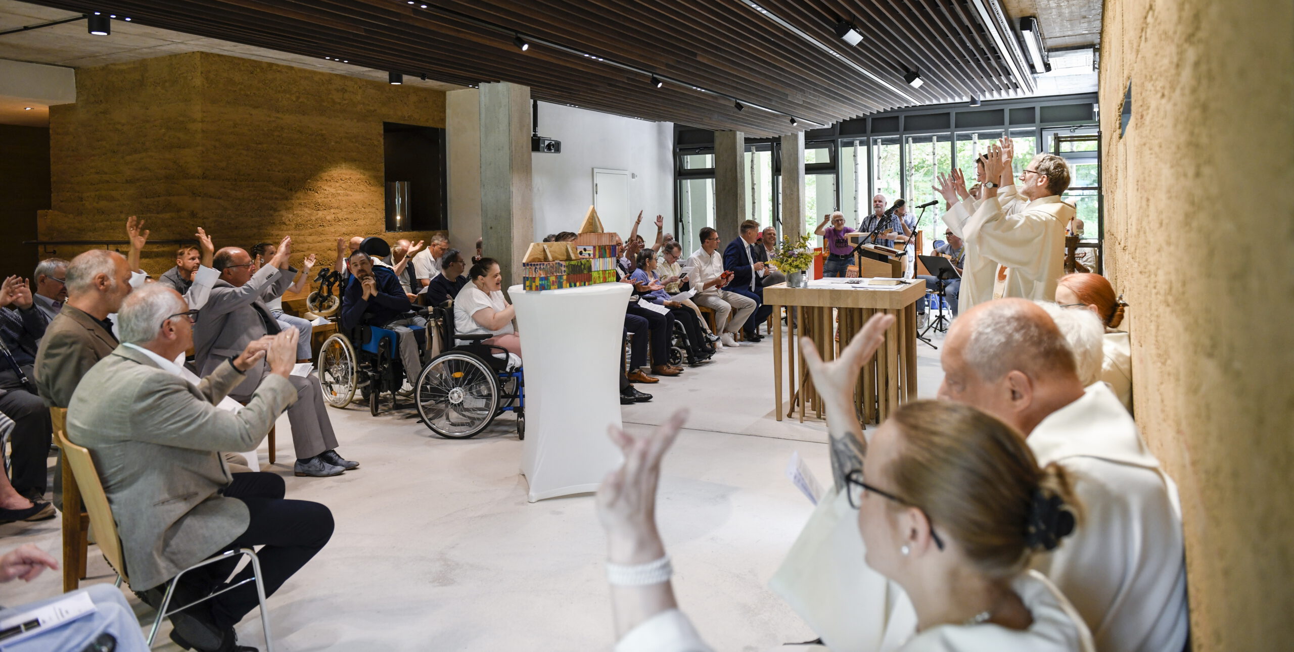 Eine Gruppe von Menschen, einige in Rollstühlen, nimmt an einem Gottesdienst in der Franziskuskapelle teil. Ein Priester an der Spitze hebt die Hände, während die Gemeinde antwortet. Das moderne Ambiente zeichnet sich durch natürliches Licht und Holzakzente aus.
