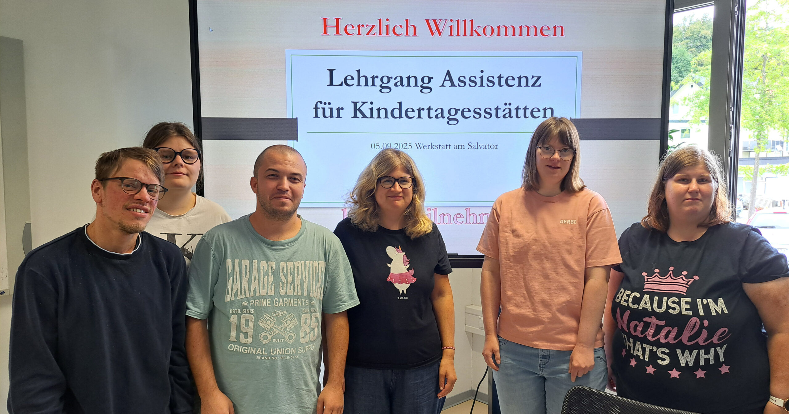 Sechs Erwachsene stehen zusammen in einem Klassenzimmer und lächeln in die Kamera. Hinter ihnen begrüßt eine Präsentationsfolie auf Deutsch die Teilnehmer des Lehrgangs für Assistent:in der Kindertagesstätte.