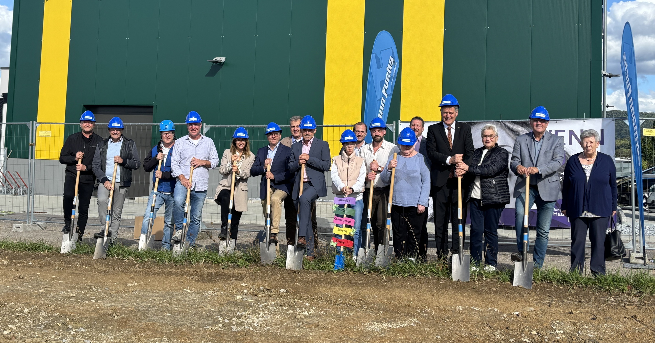 Eine Gruppe in blauen Bauhelmen steht vor dem grün-gelben Bischof-Ketteler-Haus Bargau, jeder hält eine Schaufel für den Spatenstich Bargau. Ein Lächeln und bunte Schilder zieren eine Schaufel bei dieser Zeremonie des Bischof-Ketteler-Hauses im Ersatzneubau.