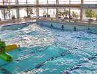 Ein Hallenbad mit klarem, blauem Wasser, sanften Wellen, ein paar Menschen, die den Pool genießen, große Fenster, die das Sonnenlicht hereinlassen, und Pflanzen an der Seite - es ist wie ein automatisch gespeicherter Entwurf von Entspannung und modernem Design.