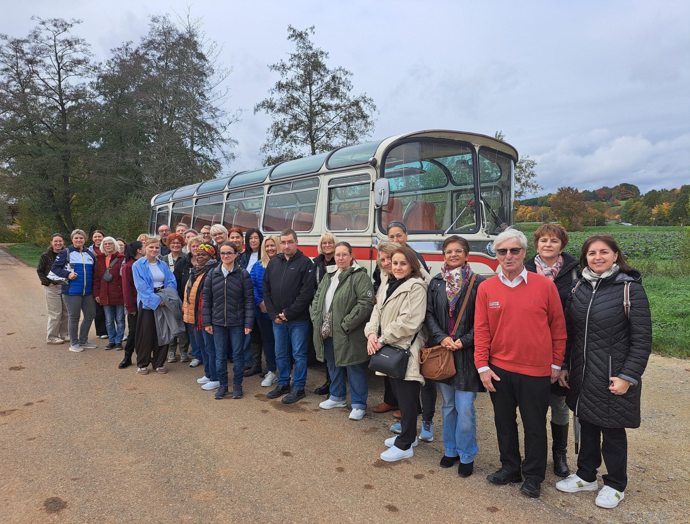 Eine Gruppe von Menschen steht an einem bewölkten Tag im Freien vor einem Oldtimerbus und posiert für ein Gruppenfoto der Jubilarfeier. Im Hintergrund sind Bäume und Grünpflanzen zu sehen, die der Altenhilfefeier eine festliche Note verleihen.