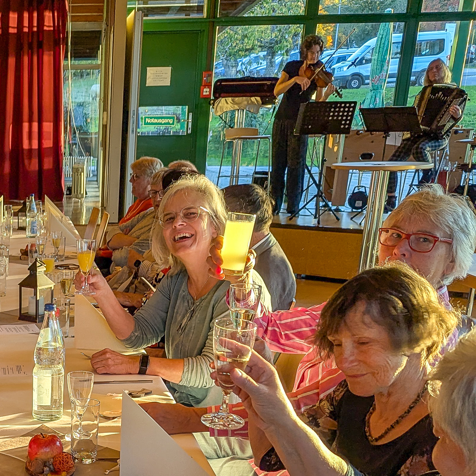 Beim Ehrenemtsfest sitzt eine Gruppe älterer Erwachsener an einer Festtafel, stößt mit Gläsern an und amüsiert sich. Auf einer kleinen Bühne spielen Musiker Geige und Akkordeon, während das Sonnenlicht durch große Fenster strömt.