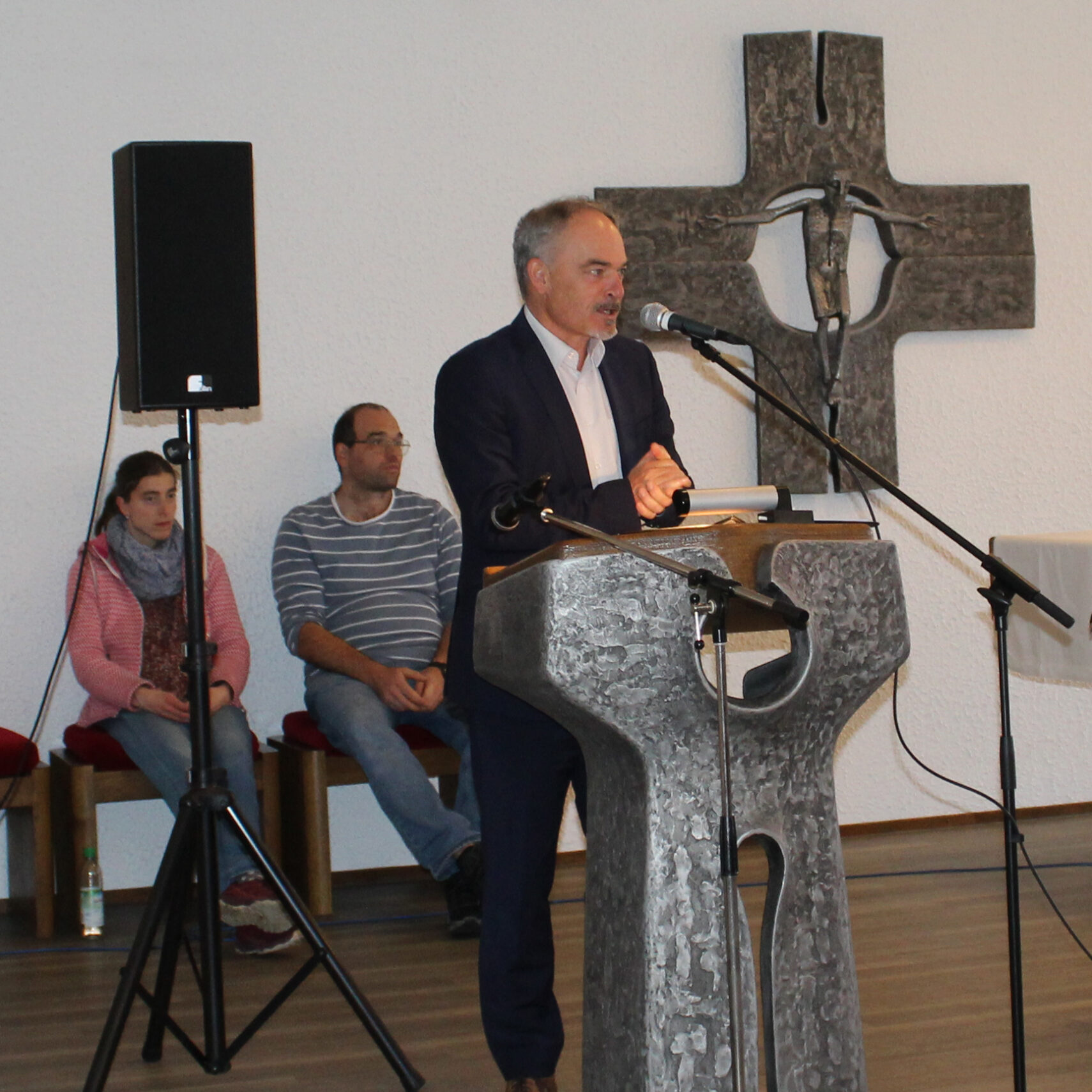 Ein Mann im Anzug spricht an einem steinernen Podium mit Mikrofon in der Kirche St. Franziskus. Zwei Personen sitzen auf einer Bank hinter ihm, während im Hintergrund ein großes Kruzifix hängt. Die Veranstaltung findet anlässlich des 30-jährigen Bestehens der Gemeinde statt.
