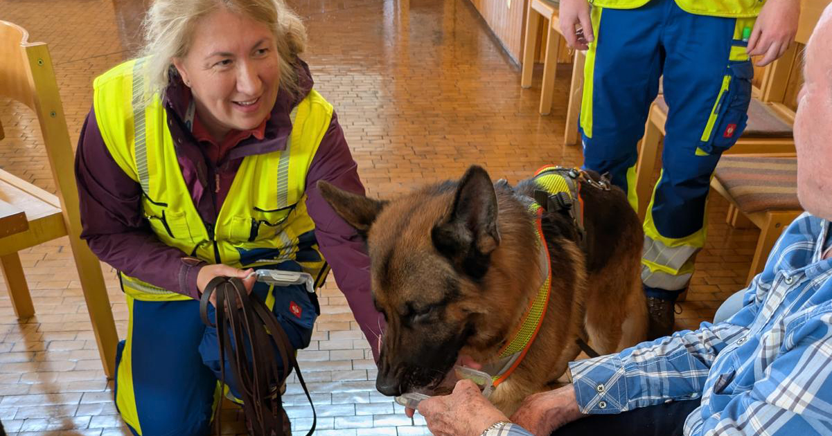 Eine Frau in einer gelben Sicherheitsweste kniet neben einem Deutschen Schäferhund, der ein Geschirr trägt, während der Hund sanft mit einem älteren sitzenden Mann in St. Elisabeth interagiert; eine weitere Person in blauen Hosen steht in der Nähe und beobachtet den Besuch der GMA. Eine Frau in einer gelben Sicherheitsweste kniet neben einem Deutschen Schäferhund, der ein Geschirr trägt, während der Hund sanft mit einem älteren sitzenden Mann in St. Elisabeth interagiert; eine weitere Person in blauen Hosen steht in der Nähe und beobachtet den Besuch der GMA.