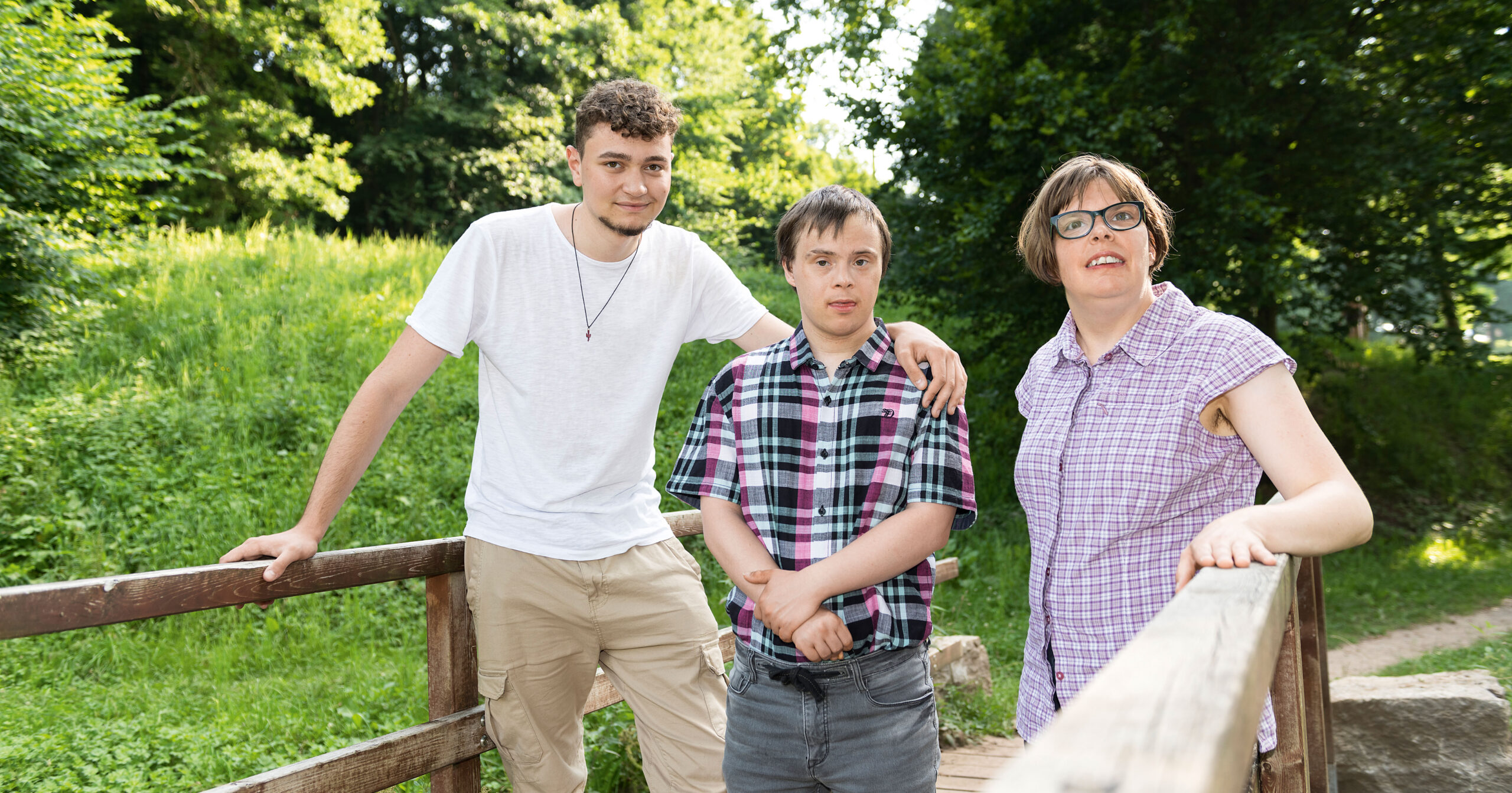 Drei Personen stehen zusammen auf einer Holzbrücke in einem Park und lächeln in die Kamera. Zwei von ihnen tragen karierte Hemden und Brillen, einer trägt ein schlichtes weißes Hemd. Grüne Bäume und Gras umgeben sie im Hintergrund. Drei Personen stehen zusammen auf einer Holzbrücke in einem Park und lächeln in die Kamera. Zwei von ihnen tragen karierte Hemden und Brillen, einer trägt ein schlichtes weißes Hemd. Grüne Bäume und Gras umgeben sie im Hintergrund.