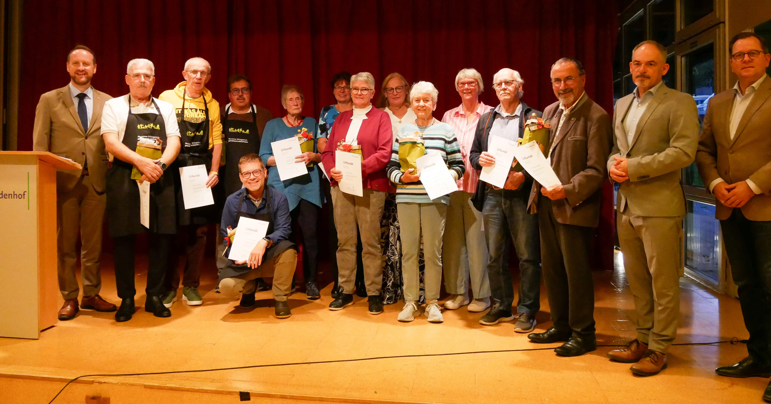 Eine Gruppe von dreizehn Personen steht auf einer Bühne beim Ehrenamtsfest, einige halten Urkunden und Blumen in der Hand und schauen in die Kamera. Fünf Männer in Anzügen stehen an den Seiten, während andere in Freizeitkleidung und Schürzen in der Mitte stehen, vor einem roten Vorhang im Hintergrund.