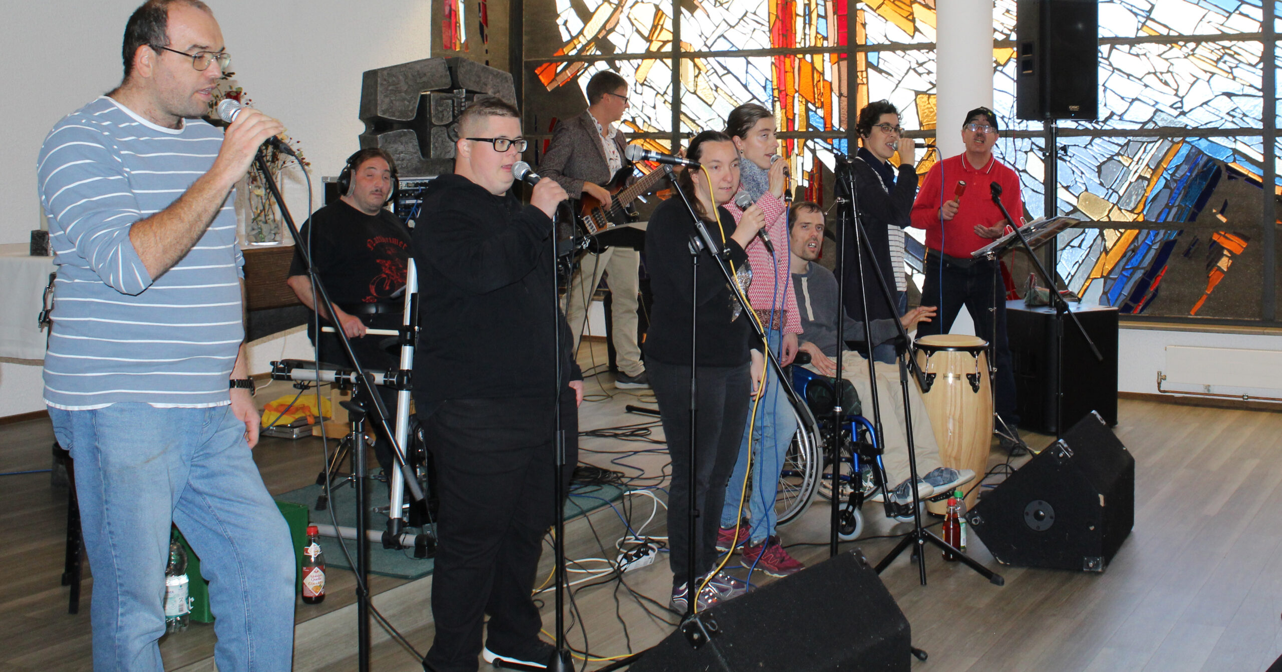 Eine Gruppe von Menschen, von denen einige im Rollstuhl sitzen, singt und spielt gemeinsam vor den bunten Glasfenstern von St. Franziskus und feiert das 30-jährige Jubiläum mit Mikrofonen, Lautsprechern und Notenständern in einem gut beleuchteten Innenraum.
