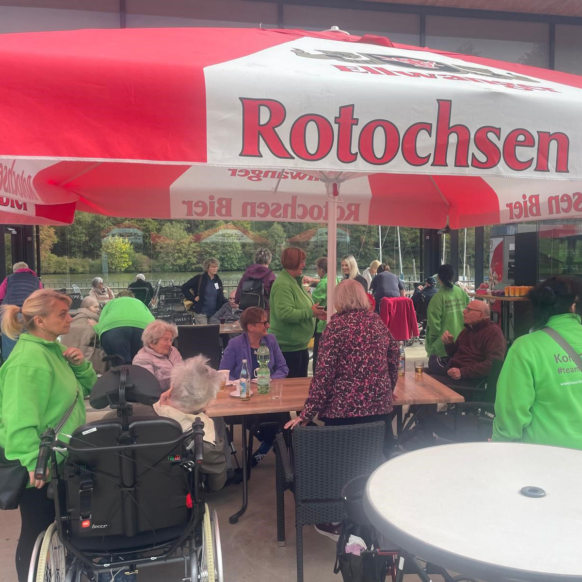 Eine Gruppe von Menschen, darunter ältere Menschen und Betreuer in grünen Jacken, sitzen und stehen um Tische im Freien unter einem großen rot-weißen Rotochsenschirm in der Nähe des Bucher Stausees. Im Hintergrund sind Bäume und der See zu sehen.