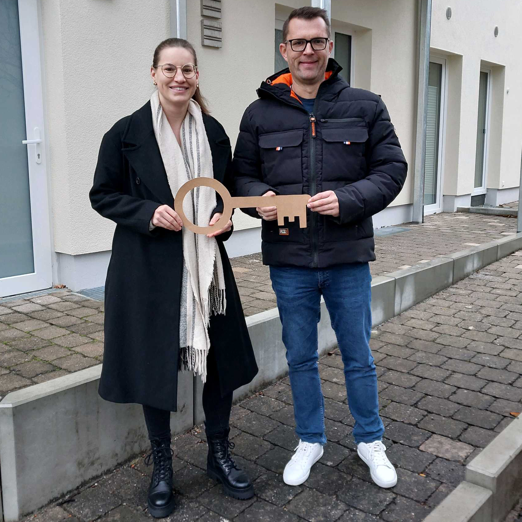Zwei Personen stehen vor einem Gebäude in der Leintalstraße, lächeln und halten einen großen symbolischen Schlüssel in der Hand. Die Frau trägt einen dunklen Mantel und ein Halstuch, der Mann eine schwarze Jacke und eine Brille. Sie feiern ihr Neues Leben in der Nähe des Eingangs.