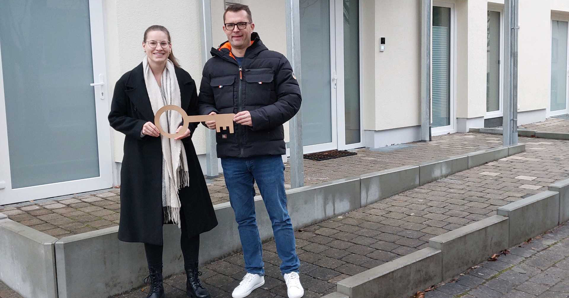 Zwei Menschen stehen vor einem Gebäude in der Leintalstraße, lächeln und halten einen großen symbolischen Schlüssel in der Hand - sie feiern den Beginn ihres neuen Lebens. Im Hintergrund sind Türen und Fenster entlang eines gepflasterten Weges zu sehen.