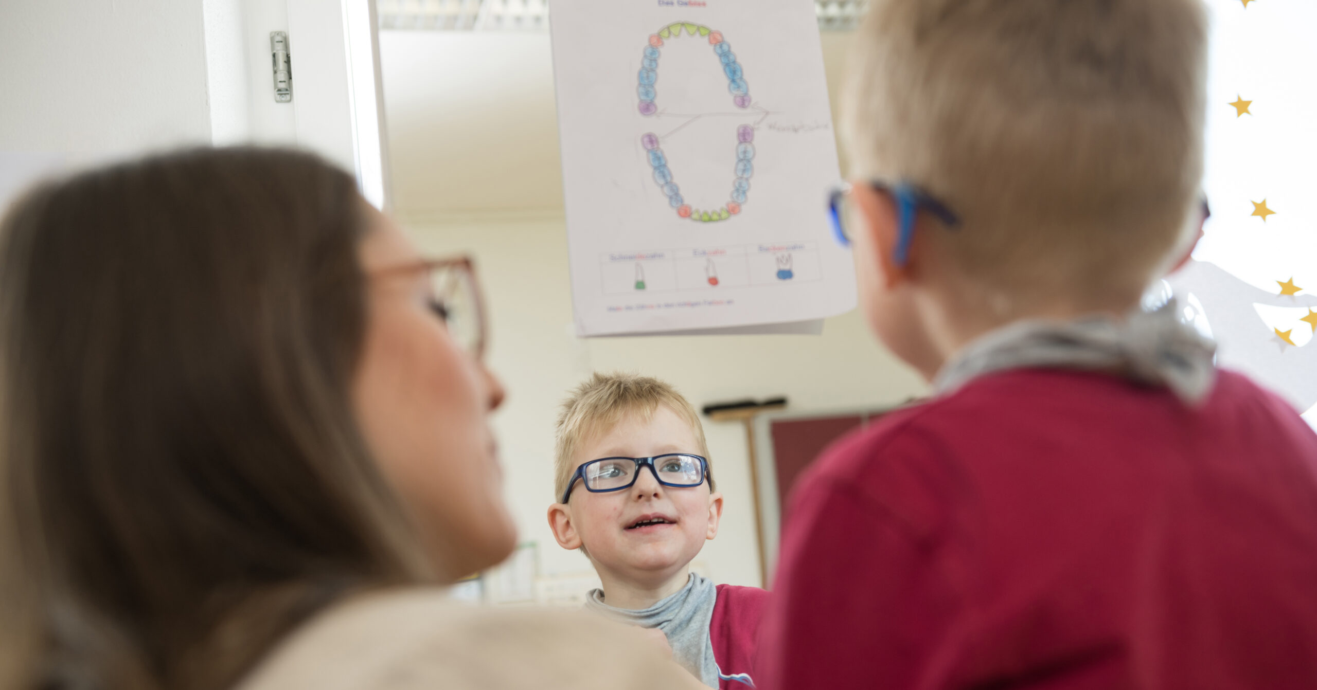 Ein kleiner Junge mit Brille betrachtet sich in einem Spiegel, während eine Frau, ebenfalls mit Brille, neben ihm sitzt. Auf dem Spiegel in diesem Neuen Schulkindergarten ist ein Diagramm der Zähne zu sehen.