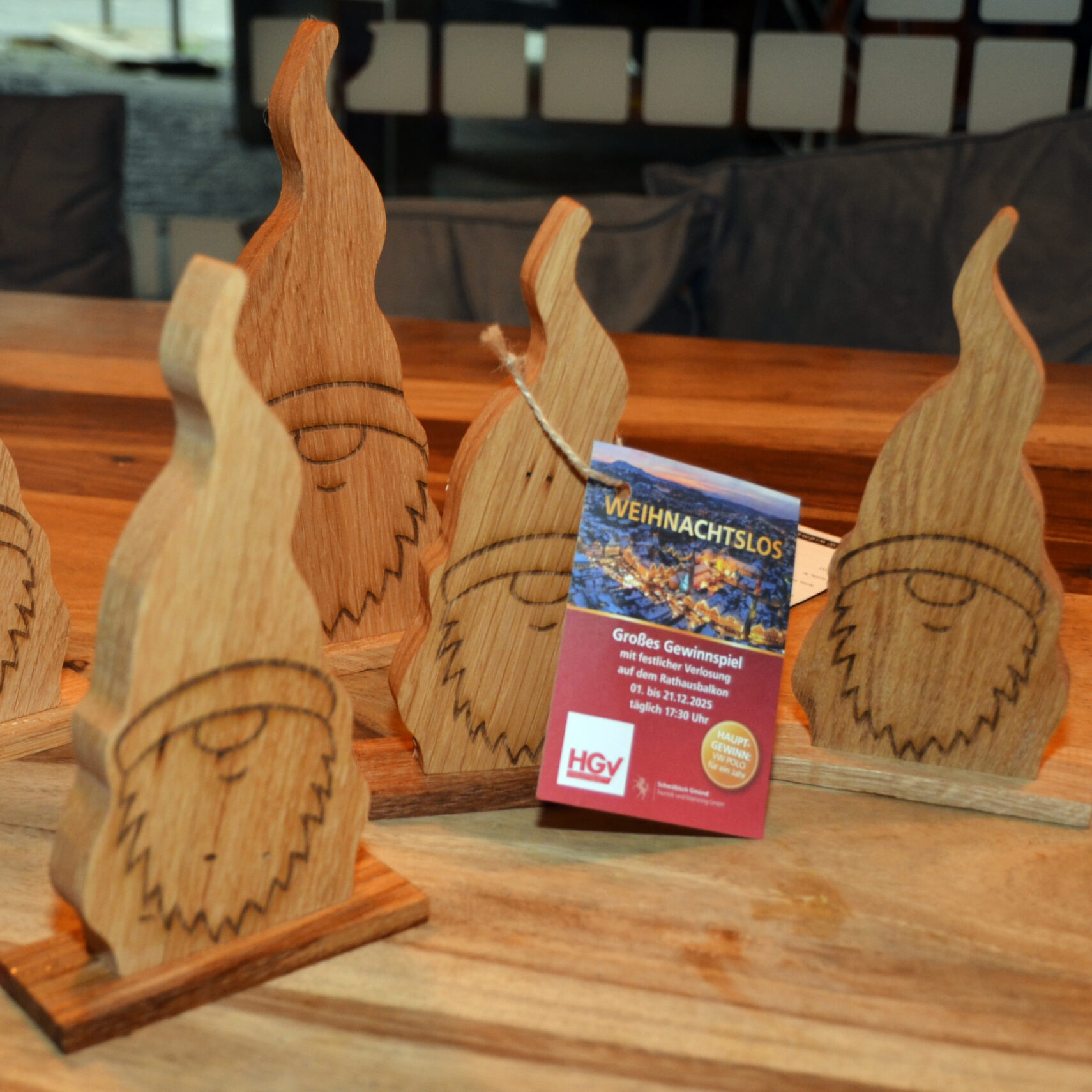 Fünf hölzerne Wichtelfiguren mit eingravierten Gesichtern sind auf einer Holzfläche angeordnet. Vor ihnen liegt ein Los mit der Aufschrift WEIHNACHTSLOS, das auf die HGV-Weihnachtsverlosung hinweist.
