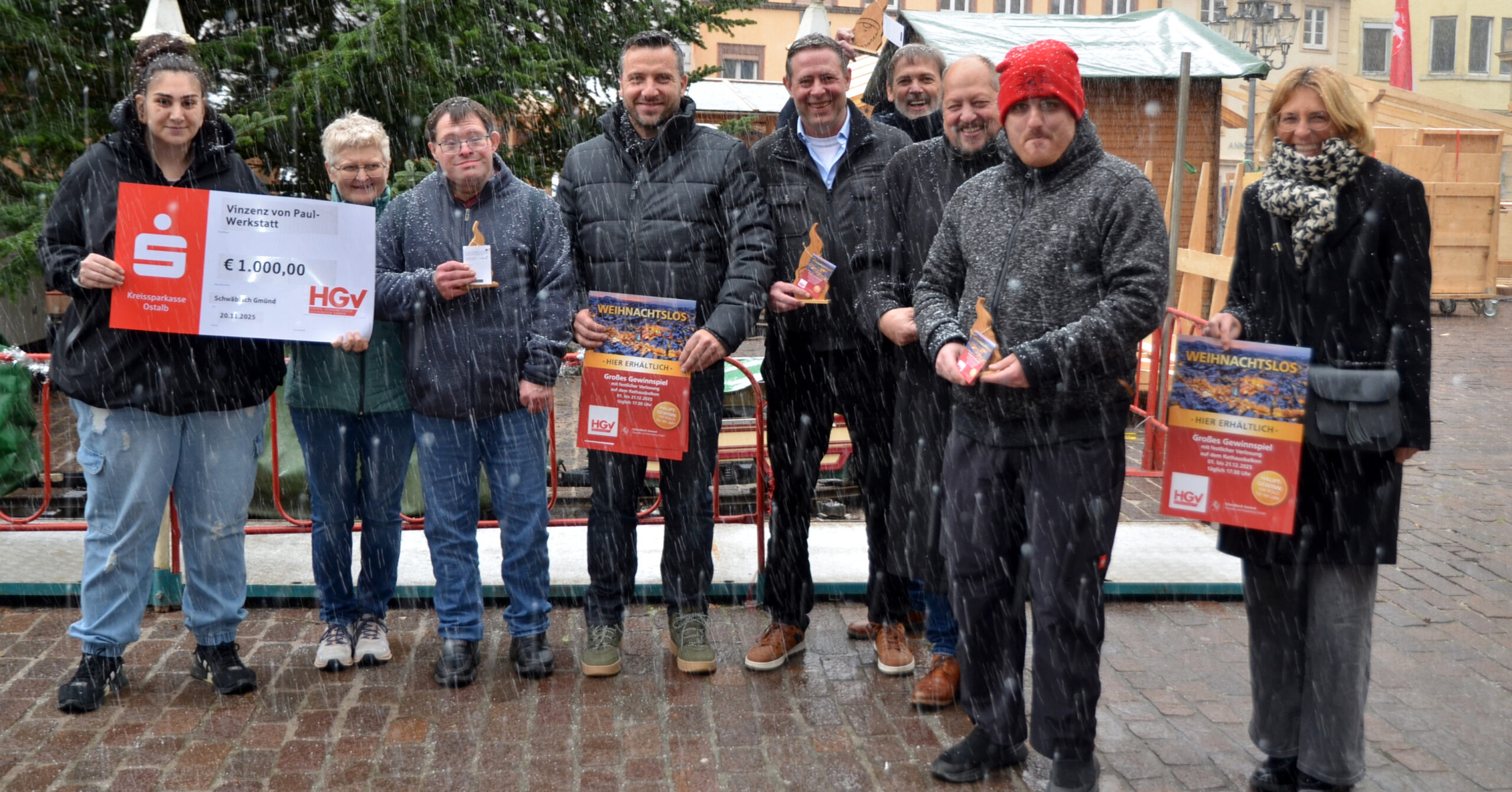 Eine Gruppe von Menschen steht während der HGV-Weihnachtsaktion im Freien im Schnee, hält Preise und einen großen Scheck über 1.000 Euro in der Hand und lächelt in die Kamera. Im Hintergrund sind ein geschmückter Weihnachtsbaum und festliche Marktstände zu sehen.