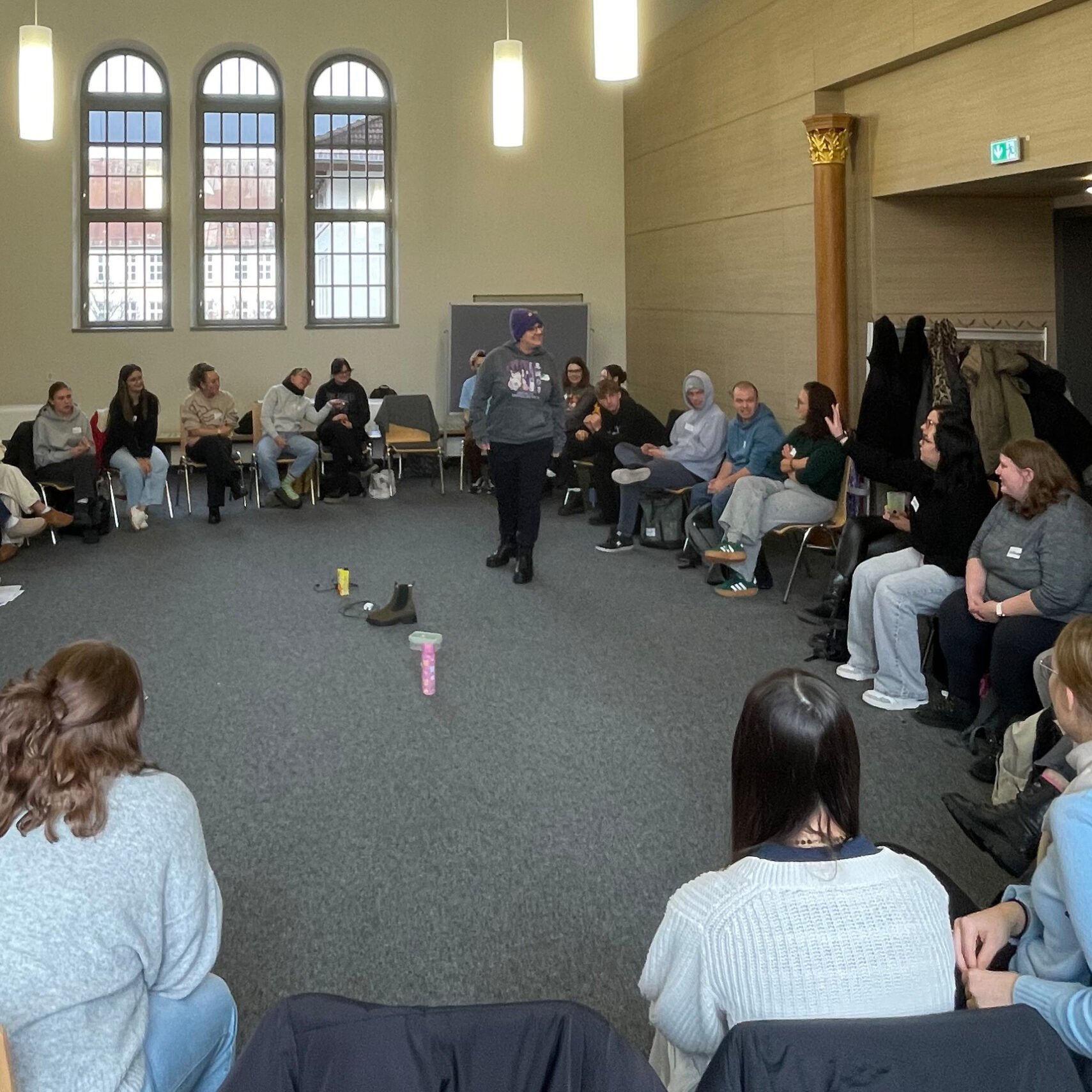 Eine Gruppe von Menschen sitzt in einem großen Kreis in einem gut beleuchteten Raum mit gewölbten Fenstern in St. Loretto. Eine Person steht in der Mitte und spricht im Rahmen einer VvP-Sitzung der Kooperation, während die anderen aufmerksam zuhören. An der Wand hängen Mäntel, auf dem Boden liegen ein paar Gegenstände.