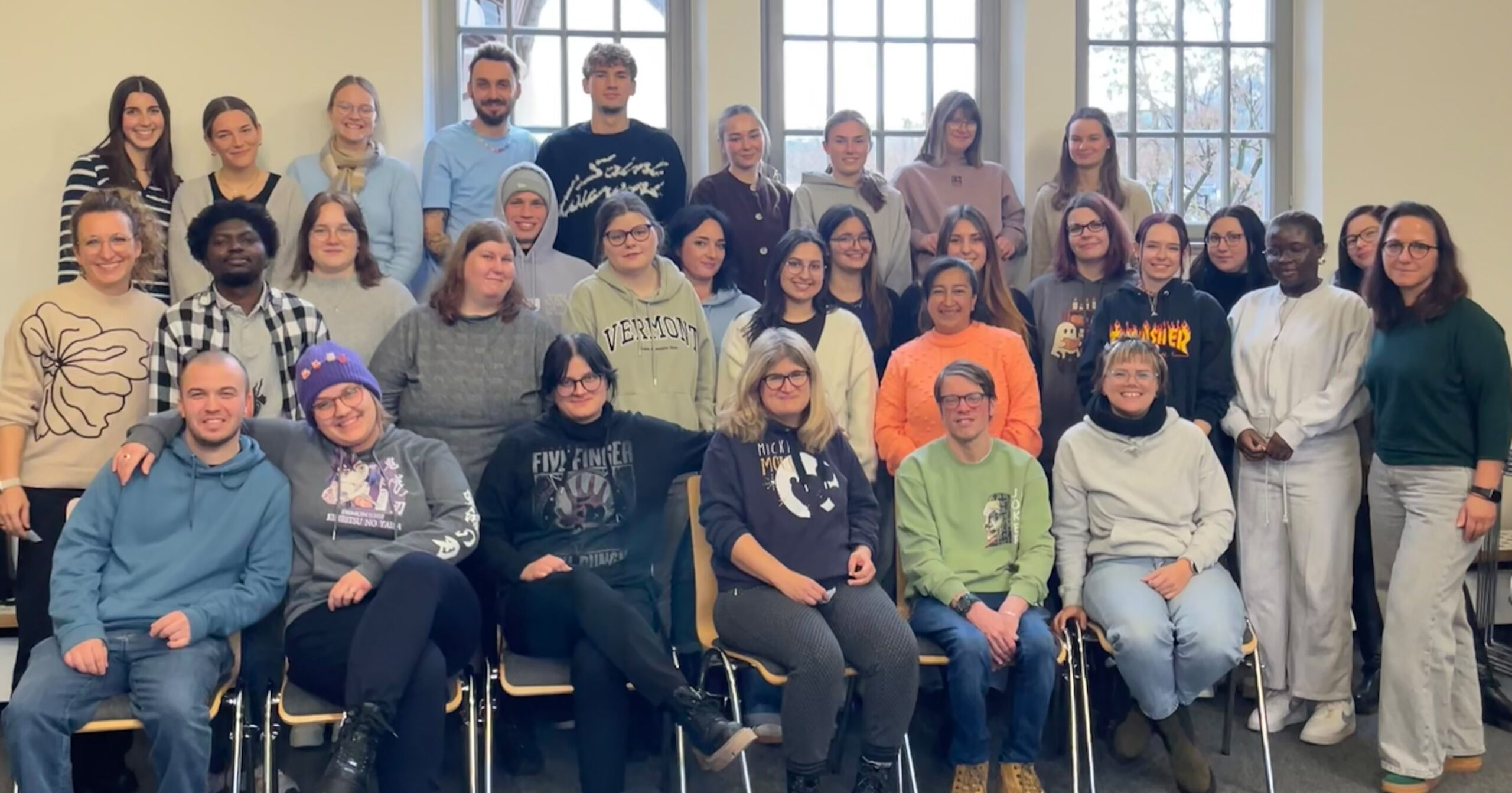 Eine Gruppe von etwa 35 Personen unterschiedlichen Alters und Hintergrunds, die zur Kooperation VvP und St. Loretto gehören, posiert drinnen vor großen Fenstern und lächelt für ein Gruppenfoto. Die meisten sitzen vorne, die anderen stehen hinten.