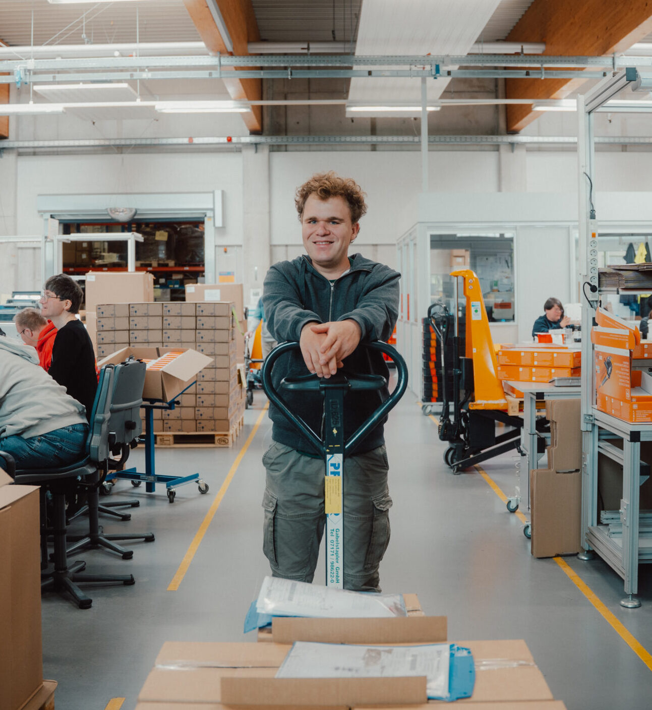 Ein Mann steht lächelnd in einem geschäftigen Lager oder einer Fabrik und stützt sich auf einen Hubwagen. Inklusionsassistenz unterstützt mehrere Arbeiter im Hintergrund, die inmitten von Kisten und Geräten Artikel zusammenstellen oder verpacken. Ein Mann steht lächelnd in einem geschäftigen Lager oder einer Fabrik und stützt sich auf einen Hubwagen. Inklusionsassistenz unterstützt mehrere Arbeiter im Hintergrund, die inmitten von Kisten und Geräten Artikel zusammenstellen oder verpacken.