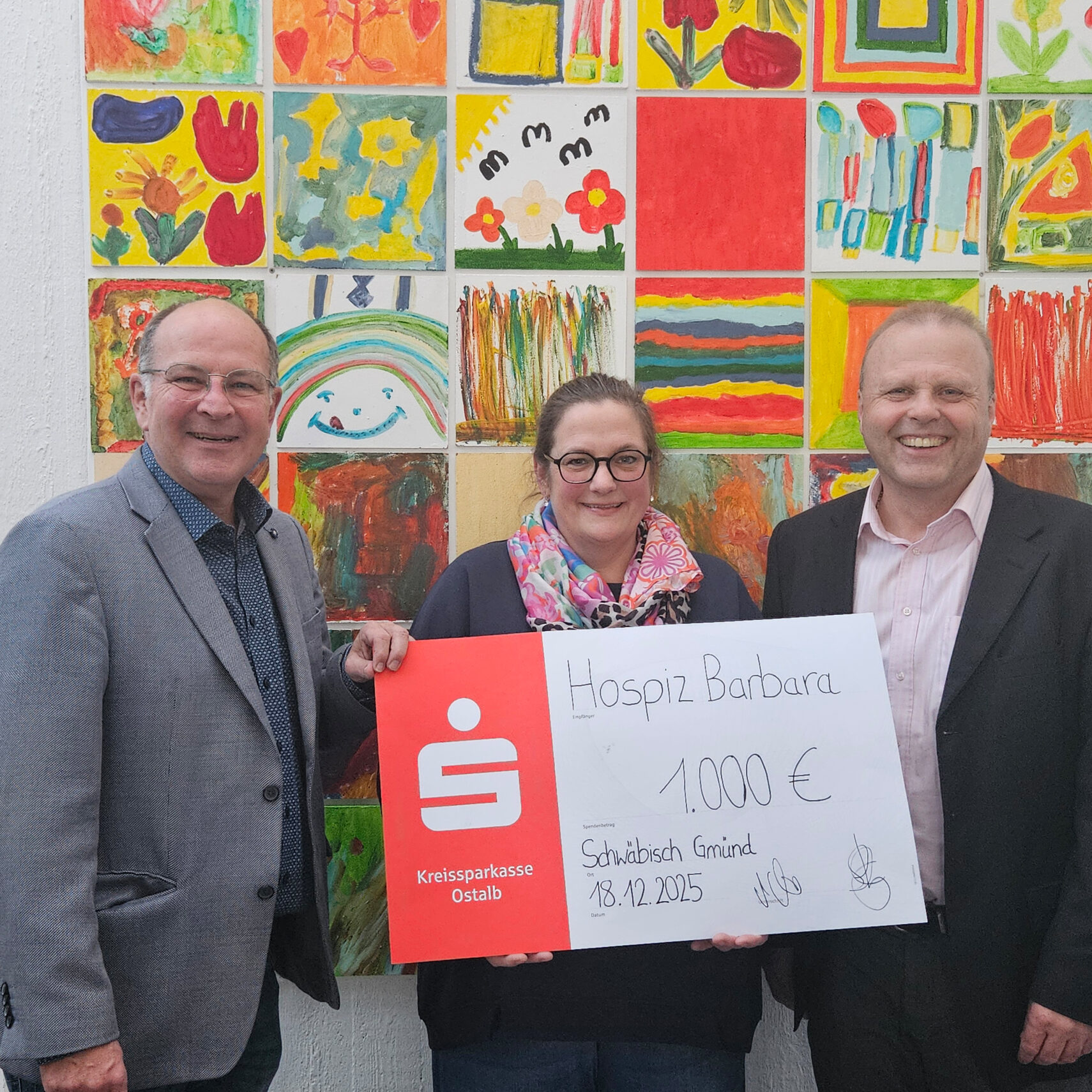 Drei lächelnde Erwachsene halten einen großen Spende-Scheck über 1.000 Euro für das Hospiz Barbara in der Hand und stehen vor einer bunten Wand mit Kinderzeichnungen. Drei lächelnde Erwachsene halten einen großen Spende-Scheck über 1.000 Euro für das Hospiz Barbara in der Hand und stehen vor einer bunten Wand mit Kinderzeichnungen.