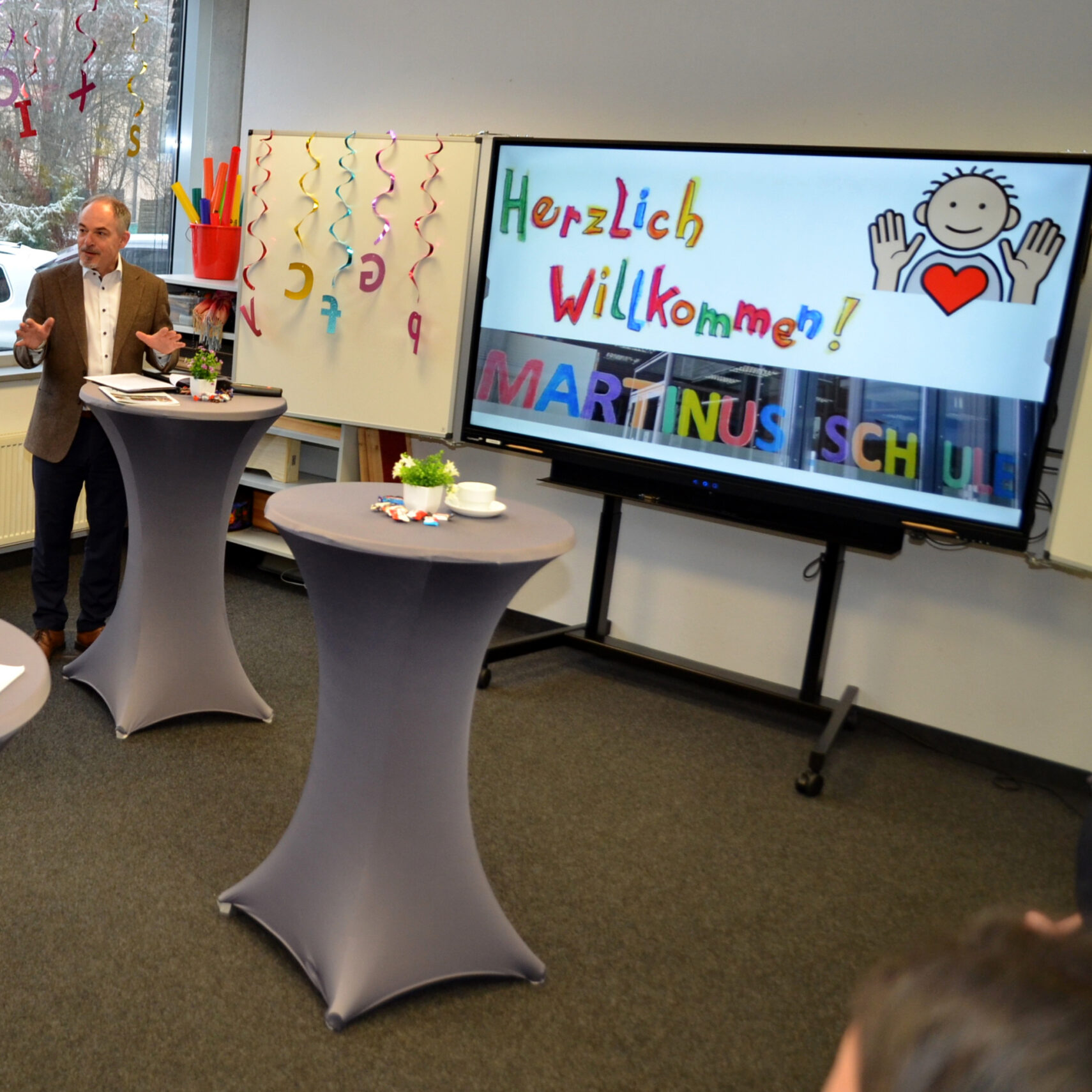 Ein Mann spricht an einem Podium in einem mit bunten Luftschlangen geschmückten Klassenzimmer während der Einweihung. Auf einem Bildschirm erscheint Herzlich Willkommen! MARTINUS SCHULE mit einer Smiley-Zeichnung, einem Herz und erhobenen Händen, um das Engagement von GmündTech zu feiern. Ein Mann spricht an einem Podium in einem mit bunten Luftschlangen geschmückten Klassenzimmer während der Einweihung. Auf einem Bildschirm erscheint Herzlich Willkommen! MARTINUS SCHULE mit einer Smiley-Zeichnung, einem Herz und erhobenen Händen, um das Engagement von GmündTech zu feiern.