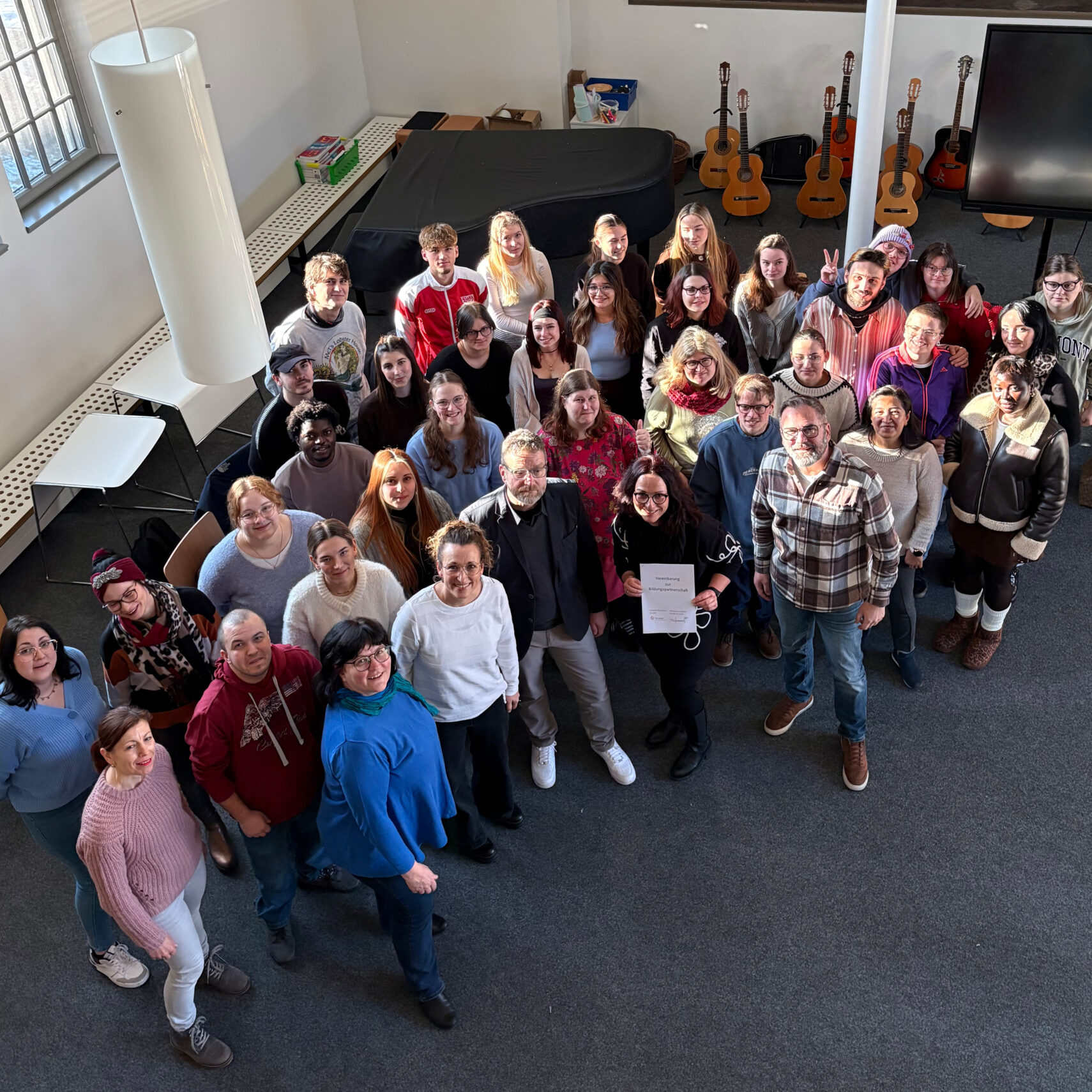 Eine Gruppe von etwa 40 Personen steht in einem hellen Raum dicht beieinander, lächelt und blickt in die Kamera. Im Hintergrund sind mehrere Gitarren zu sehen, dazu Bänke und große Fenster – ein lebendiger Moment in der Kooperation St. Loreto. Eine Gruppe von etwa 40 Personen steht in einem hellen Raum dicht beieinander, lächelt und blickt in die Kamera. Im Hintergrund sind mehrere Gitarren zu sehen, dazu Bänke und große Fenster - ein lebendiger Moment in der Kooperation St. Loreto.