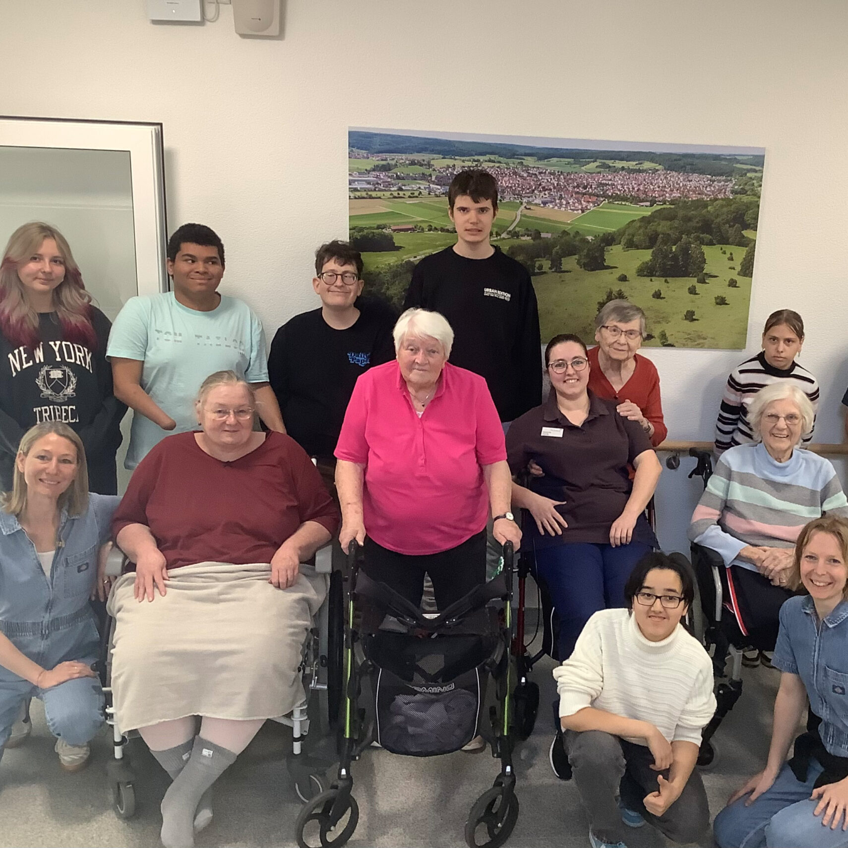 Eine Gruppe älterer Erwachsener und jüngerer Menschen posiert und lächelt gemeinsam in St. Georg, wo sie sich für ein Kunstprojekt vor einem großen Landschaftsfoto an der Wand versammelt haben. Einige sitzen, andere stehen, und einige nutzen Mobilitätshilfen in dieser einladenden Innenumgebung. Eine Gruppe älterer Erwachsener und jüngerer Menschen posiert und lächelt gemeinsam in St. Georg, wo sie sich für ein Kunstprojekt vor einem großen Landschaftsfoto an der Wand versammelt haben. Einige sitzen, andere stehen, und einige nutzen Mobilitätshilfen in dieser einladenden Innenumgebung.