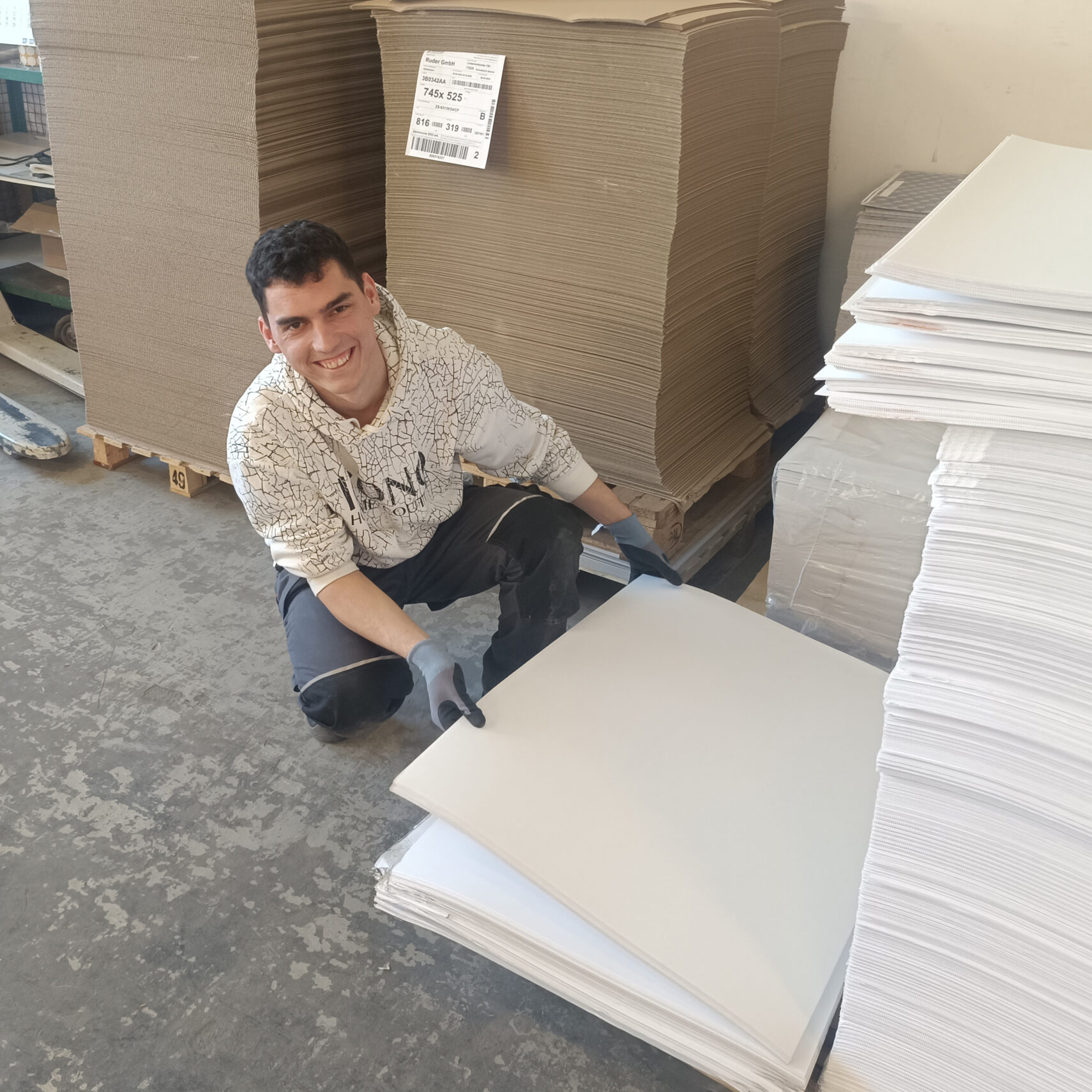Eine Person mit Handschuhen und weißem Kapuzenpulli kniet neben großen Stapeln von Pappe und weißen Papierbögen in einer Industrieumgebung und lächelt in die Kamera – ein praktisches Praktikum in den Job. Eine Person mit Handschuhen und weißem Kapuzenpulli kniet neben großen Stapeln von Pappe und weißen Papierbögen in einer Industrieumgebung und lächelt in die Kamera - ein praktisches Praktikum in den Job.