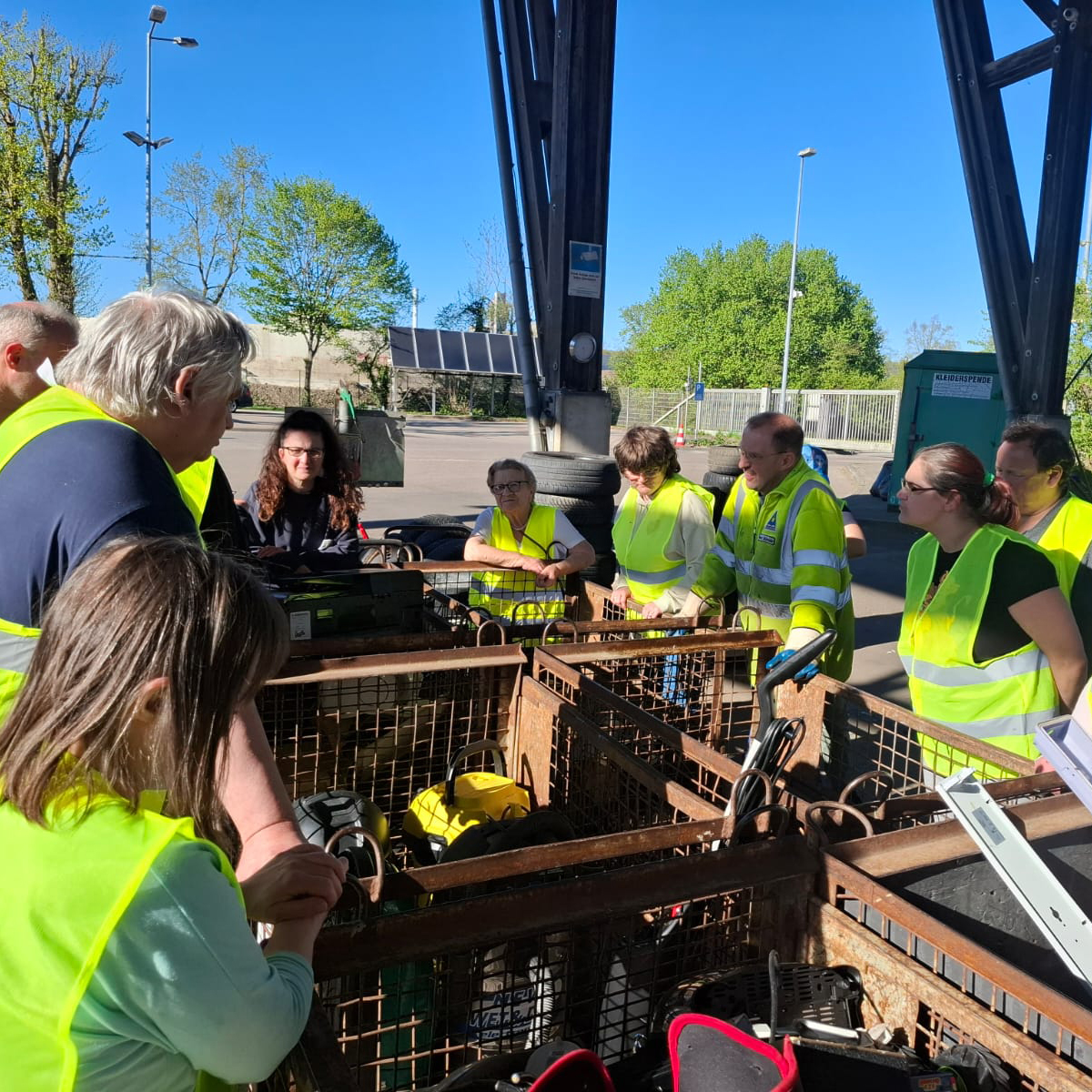 Eine Gruppe von Menschen mit gelben Sicherheitswesten steht um Metalltonnen mit elektronischen Geräten im Freien am Haus der Teilhabe Göppingen und hört einem Vertreter der Kreisabfallwirtschaft zu, der etwas erklärt. Im Hintergrund sind Bäume und ein Zaun zu sehen. Eine Gruppe von Menschen mit gelben Sicherheitswesten steht um Metalltonnen mit elektronischen Geräten im Freien am Haus der Teilhabe Göppingen und hört einem Vertreter der Kreisabfallwirtschaft zu, der etwas erklärt. Im Hintergrund sind Bäume und ein Zaun zu sehen.