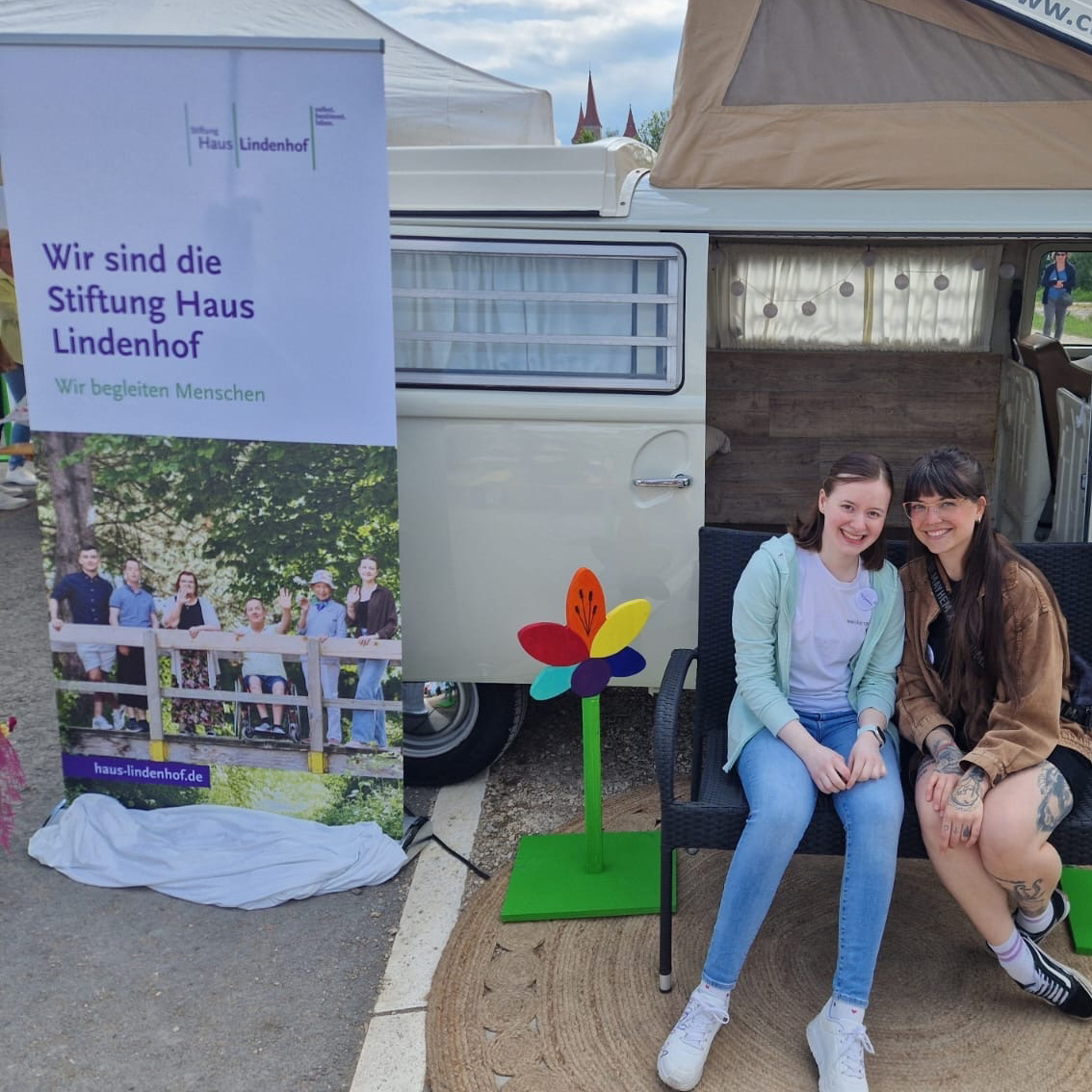 Zwei junge Frauen sitzen lächelnd auf einer Bank vor einem weißen Lieferwagen und bunten Blumen auf der Landesgartenschau. Auf einem Transparent neben ihnen steht „Wir sind die Stiftung Haus Lindenhof – Wir begleiten Menschen“, darunter ein Gruppenfoto. Zwei junge Frauen sitzen lächelnd auf einer Bank vor einem weißen Lieferwagen und bunten Blumen auf der Landesgartenschau. Auf einem Transparent neben ihnen steht