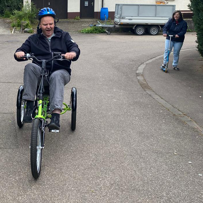 Ein Mann mit blauem Helm fährt auf einem asphaltierten Weg in Wäschenbeuren mit einem grünen Dreirad, während eine Frau in dunkler Jacke und Jeans auf einem Roller hinter ihm herfährt. Im Hintergrund sind ein Anhänger und ein Gebäude zu sehen. Ein Mann mit blauem Helm fährt auf einem asphaltierten Weg in Wäschenbeuren mit einem grünen Dreirad, während eine Frau in dunkler Jacke und Jeans auf einem Roller hinter ihm herfährt. Im Hintergrund sind ein Anhänger und ein Gebäude zu sehen.